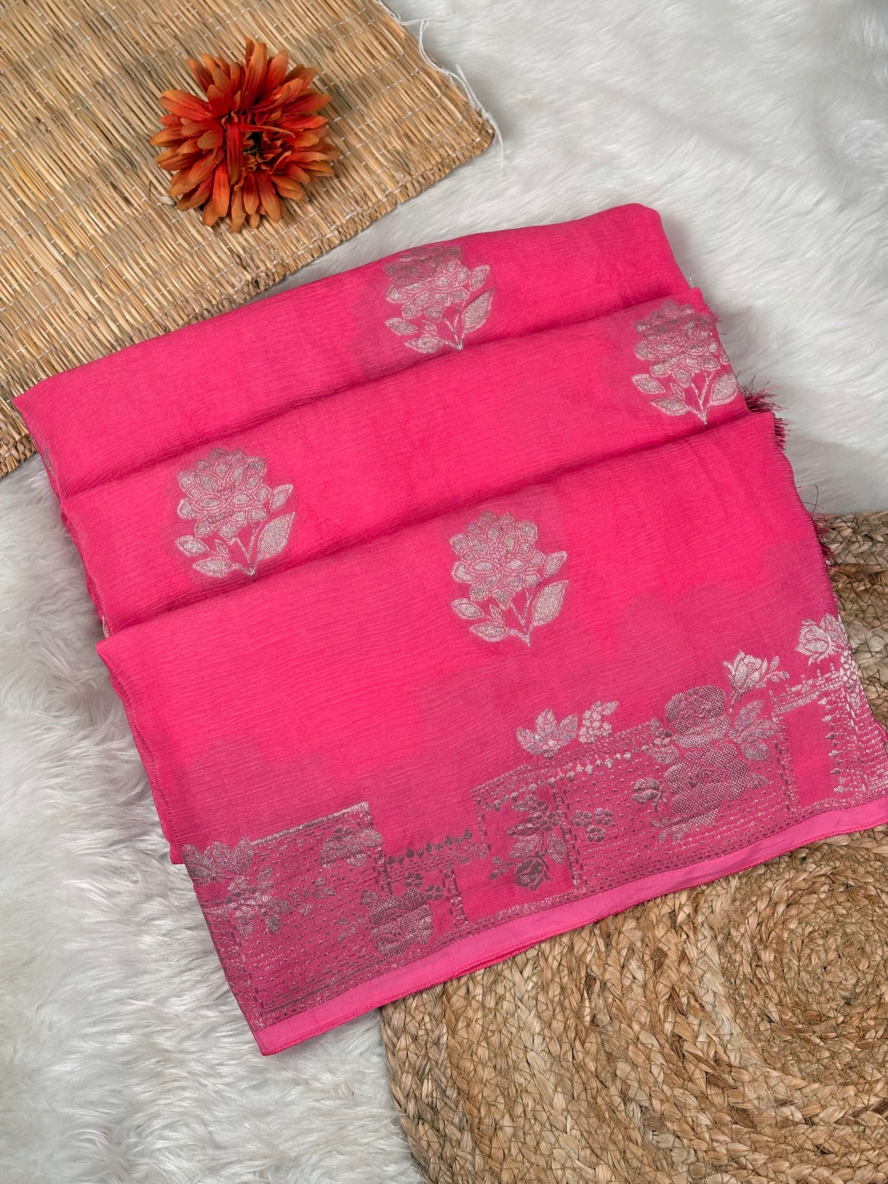 A folded bright pink saree with light pink floral motifs and a decorative border, styled on a white faux-fur surface with woven mat props and a small orange flower.