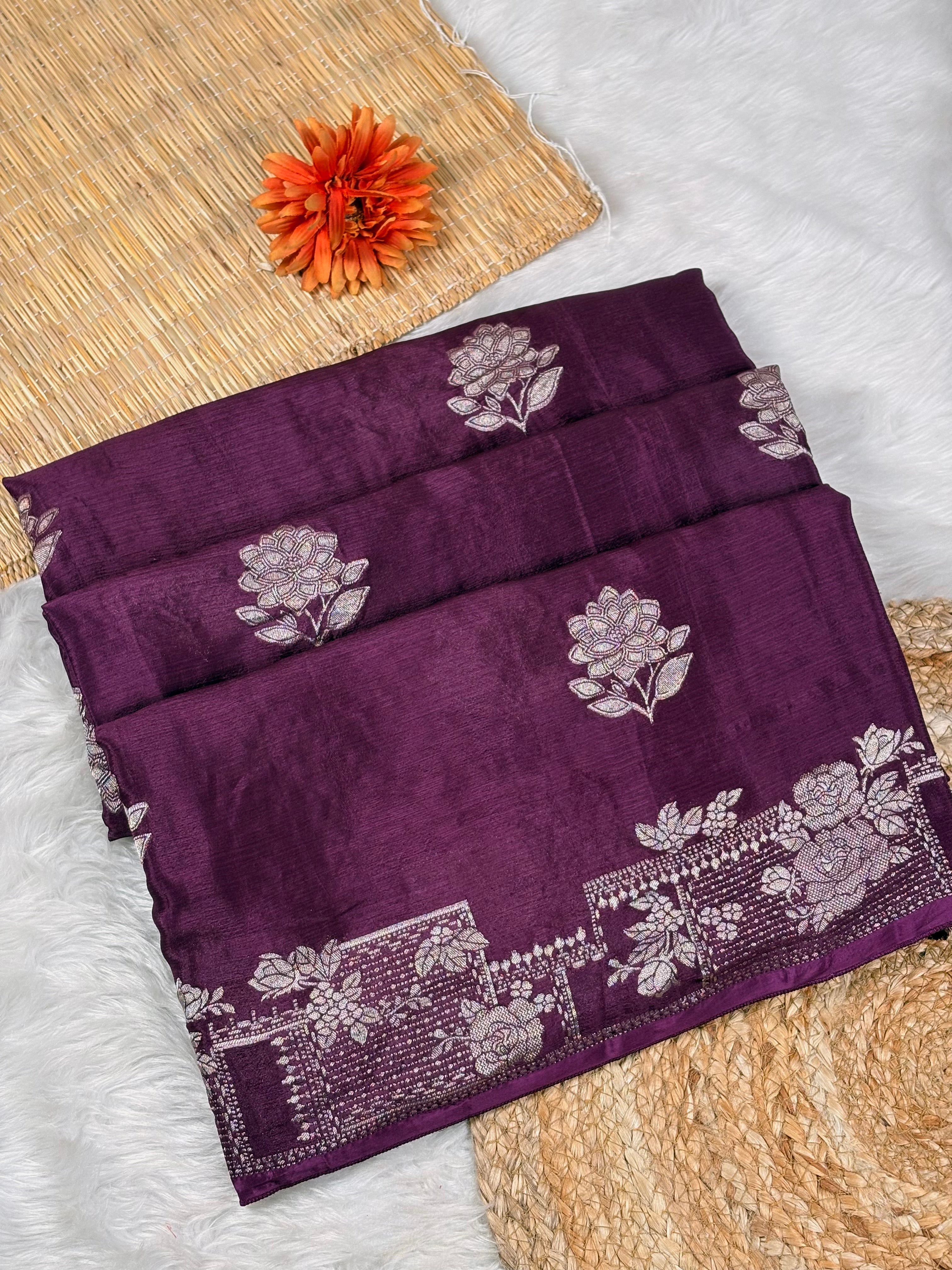 A folded deep purple silk saree with intricate water gold floral and border motifs, displayed on a white faux fur surface with a woven mat and orange flower prop.