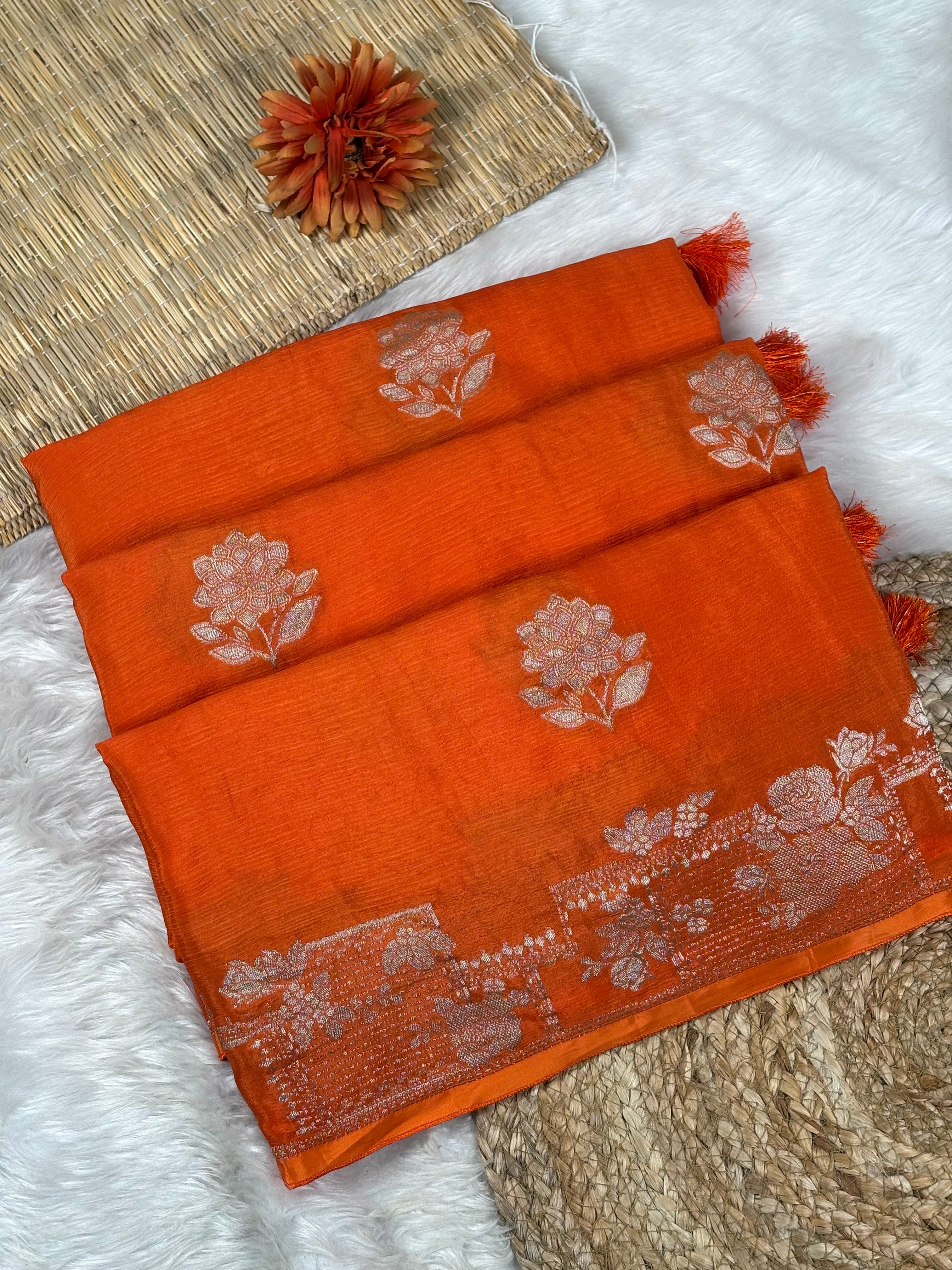 An orange woven saree with water gold floral motifs and a decorative border, styled on a white faux fur surface with a woven mat and a dried flower.