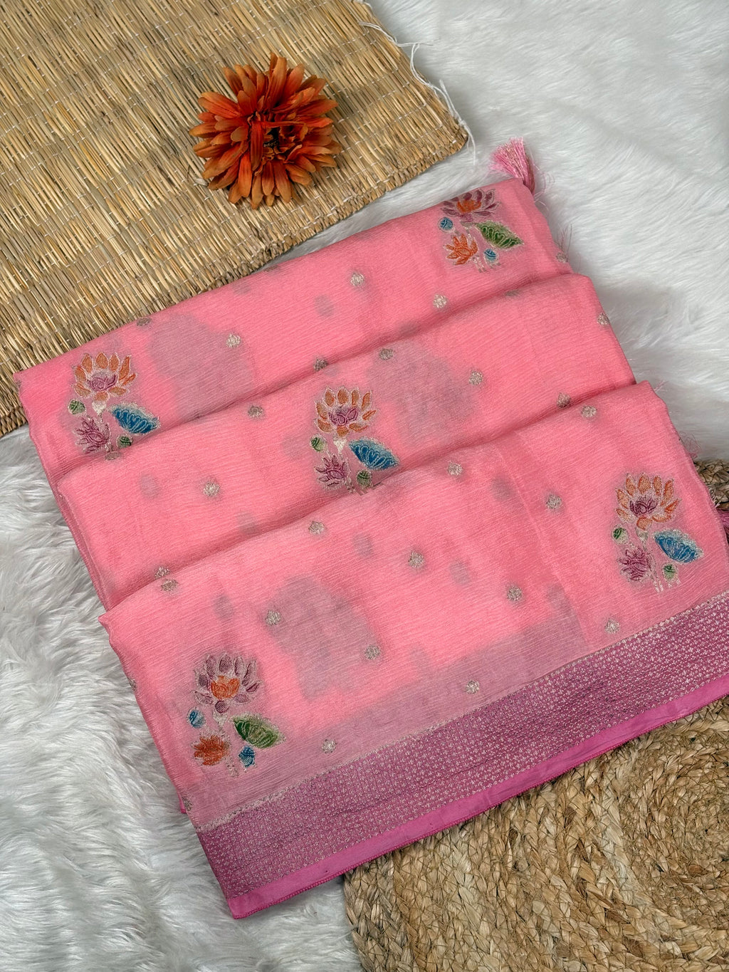 A folded pink saree with floral weaving and small metallic dot motifs, displayed on a white faux-fur surface with a woven mat and a decorative orange flower prop.