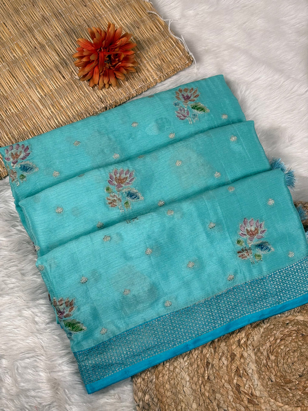 A folded turquoise blue saree with floral weaving and metallic motifs, styled on a white faux-fur surface with a woven mat and a decorative orange flower.