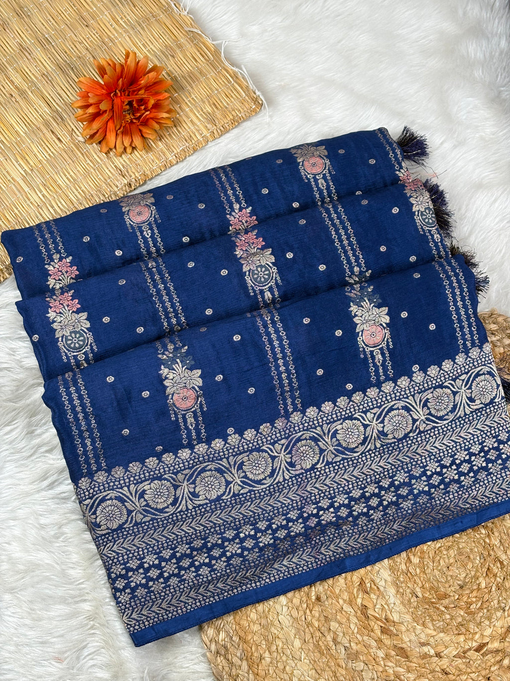 A folded navy blue saree with intricate silver zari weaving, floral motifs, and a decorative border, styled on a white faux fur surface with a woven mat and an orange flower accent.
