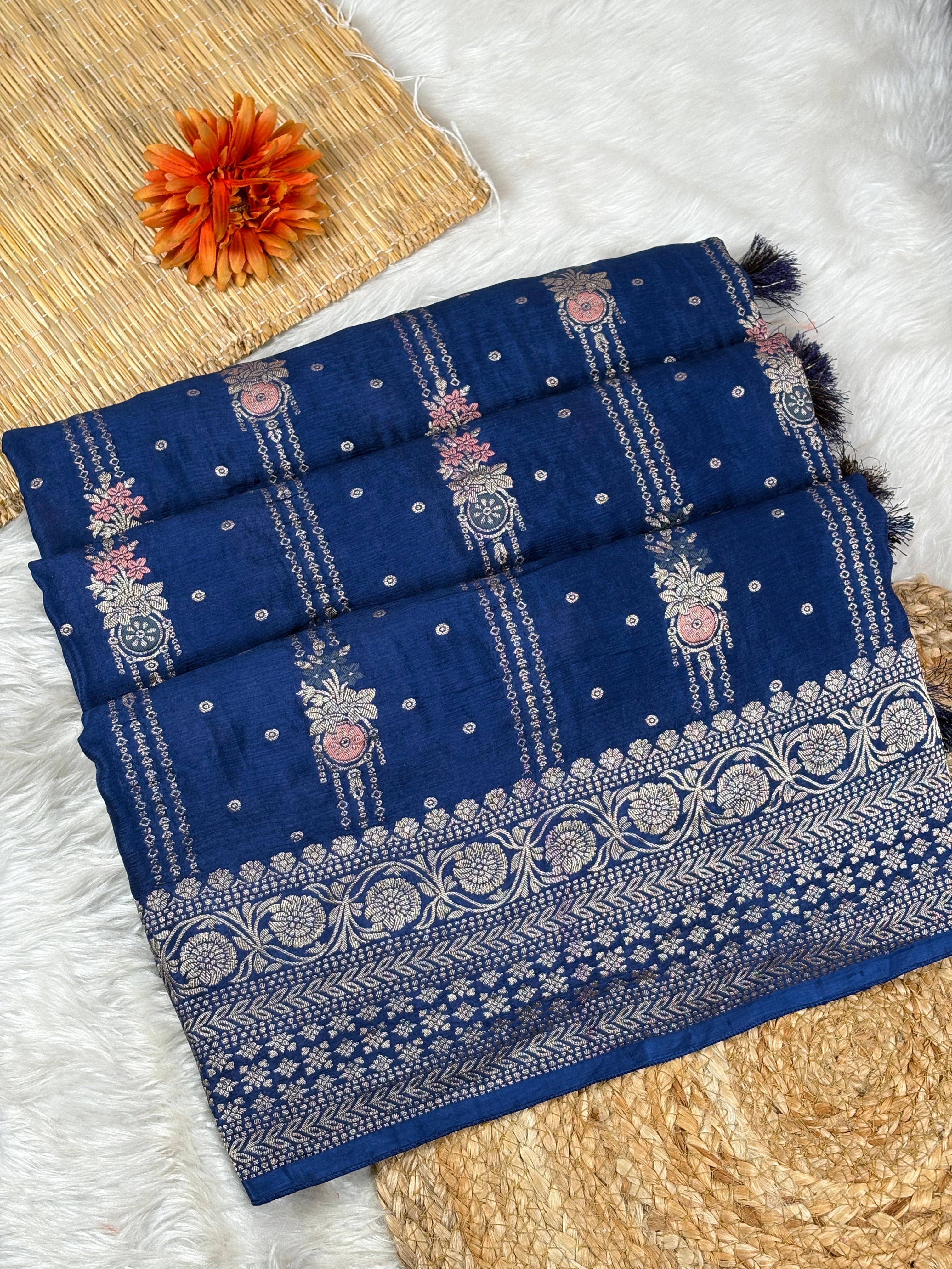 A folded navy blue saree with intricate silver zari weaving, floral motifs, and a decorative border, styled on a white faux fur surface with a woven mat and an orange flower accent.