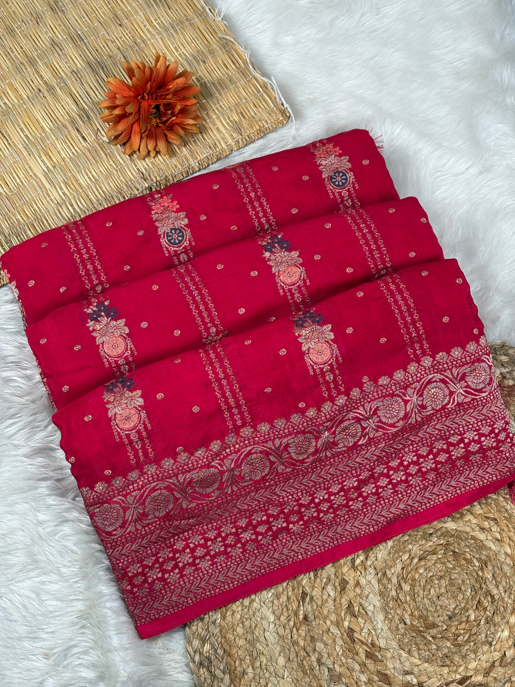 A folded red Banarasi silk saree with intricate gold and silver zari weaving, displayed on a styled background of white faux fur, a woven mat, and a small orange flower.