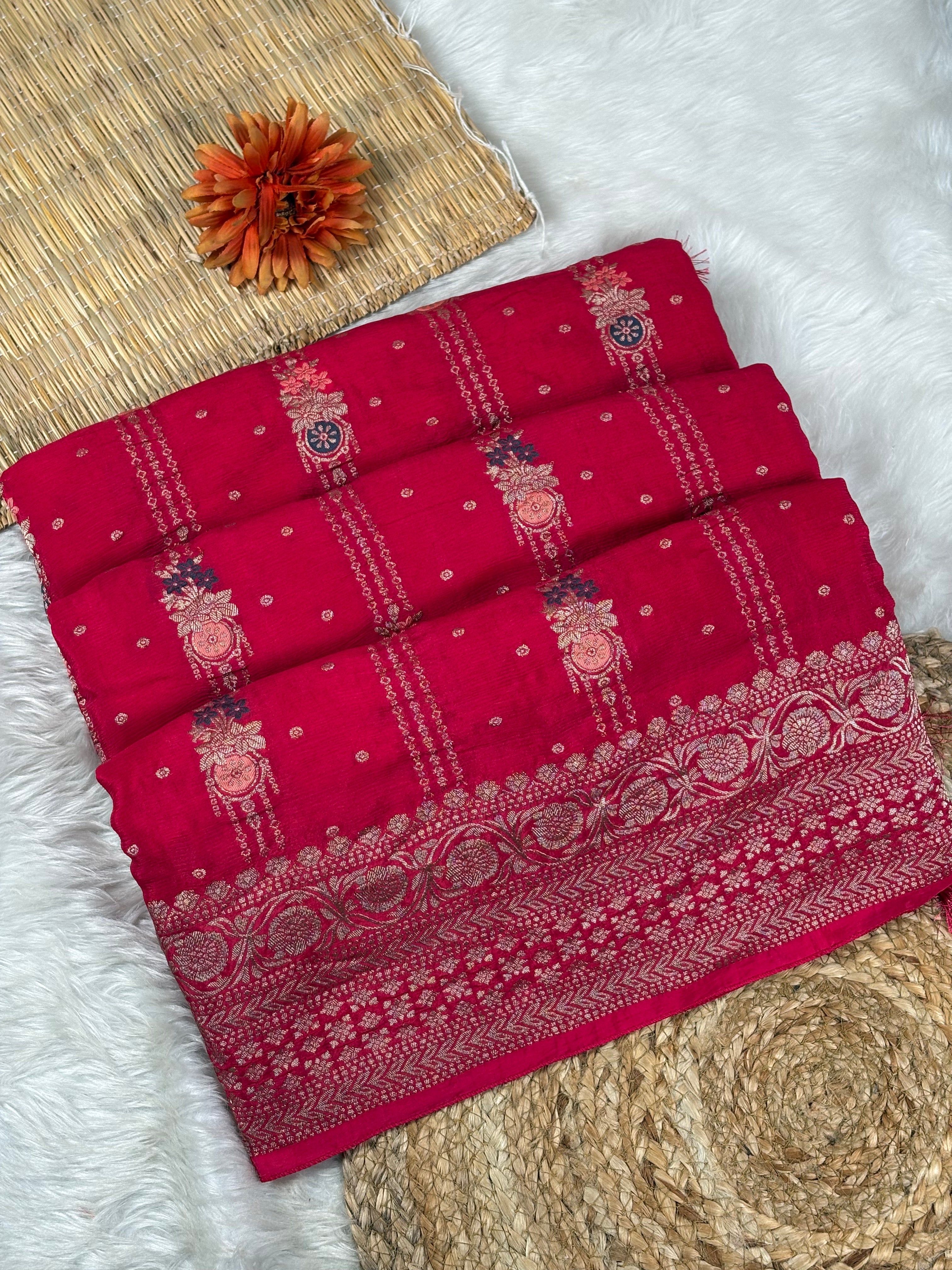 A folded red Banarasi silk saree with intricate gold and silver zari weaving, displayed on a styled background of white faux fur, a woven mat, and a small orange flower.