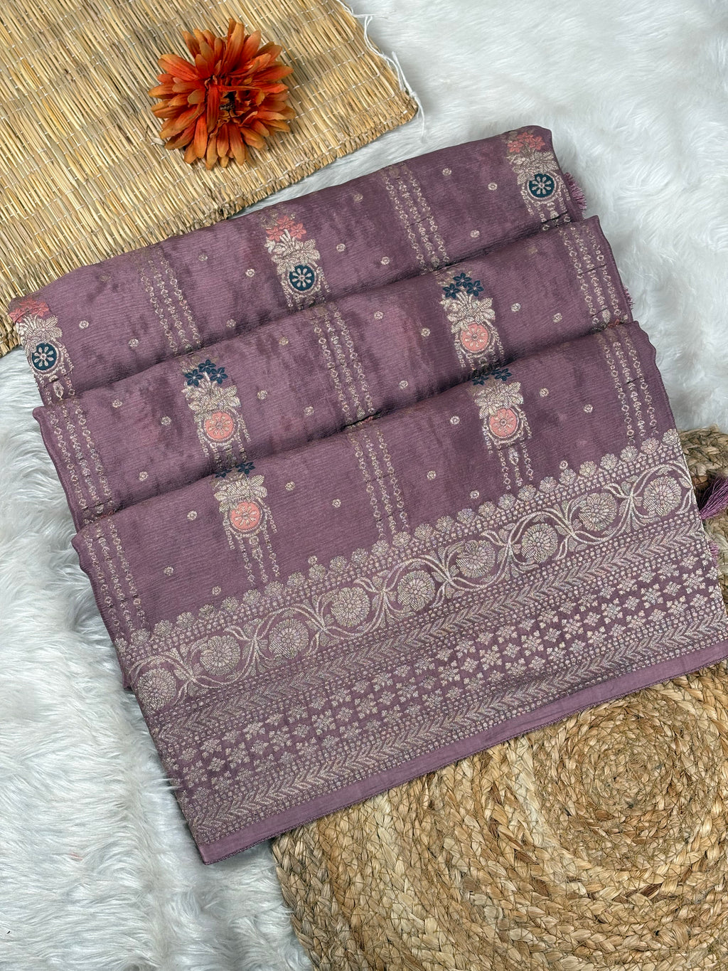 A folded dusty purple saree with intricate woven patterns and borders, styled on a white faux fur surface with a woven mat and a decorative orange flower.