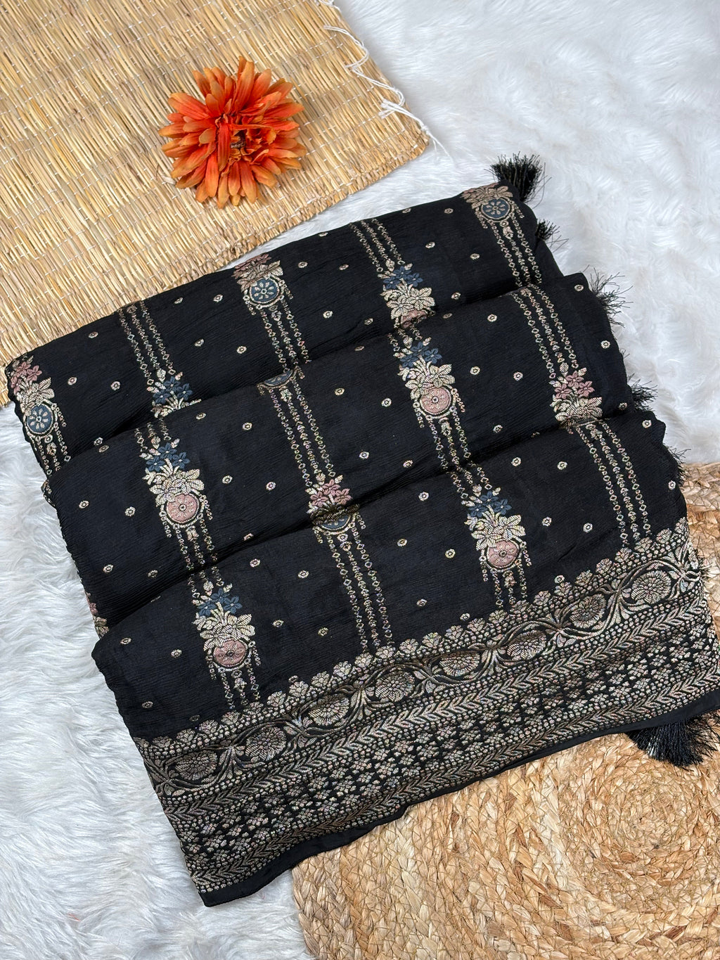 A folded black saree with intricate gold, pink, and blue floral and geometric weaving, displayed on a white faux fur surface with a woven mat and an orange flower as props.