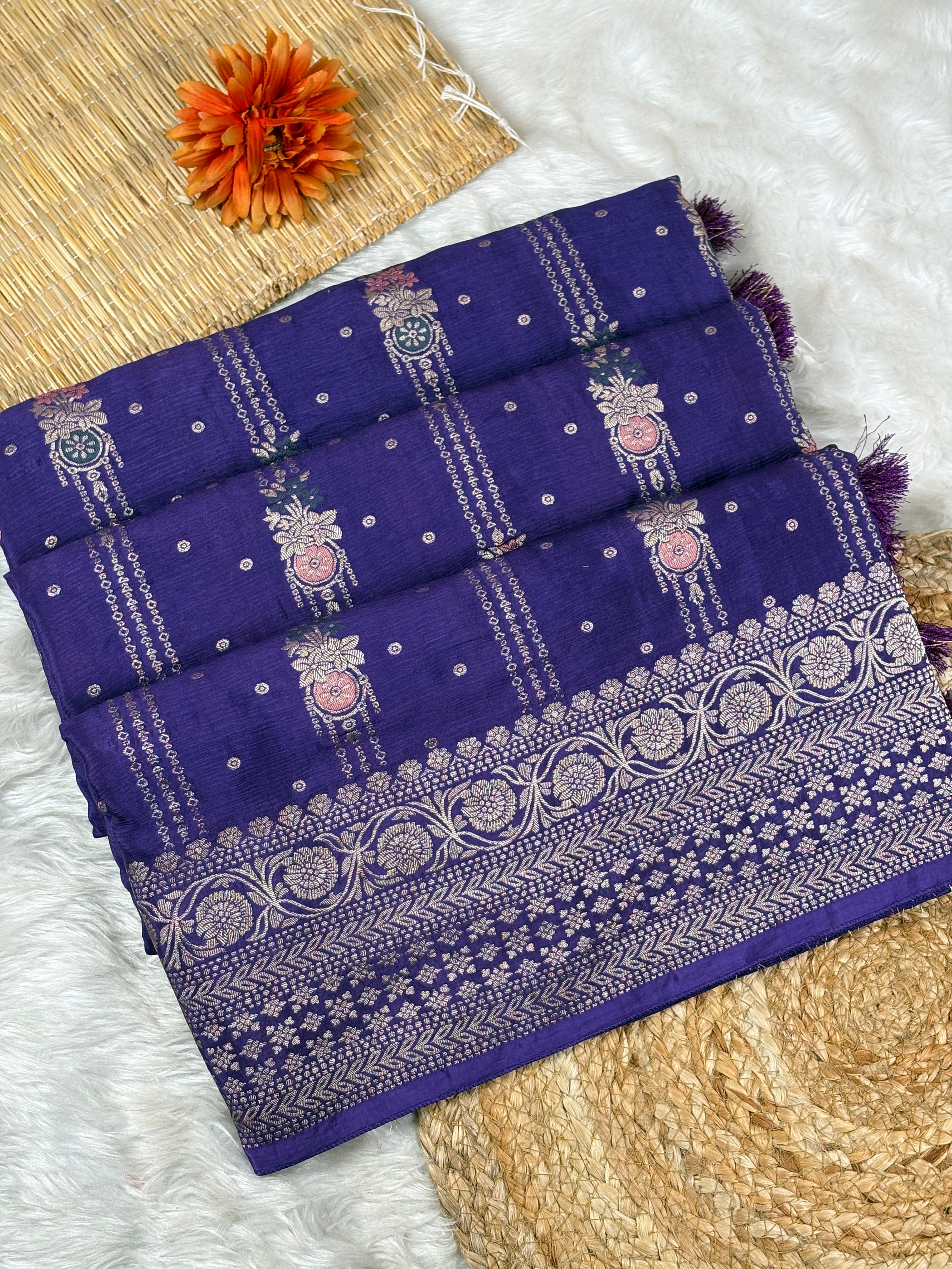 A folded purple Banarasi silk saree with intricate silver zari weaving, displayed on a white faux fur rug alongside a straw mat, a woven mat, and an orange flower.