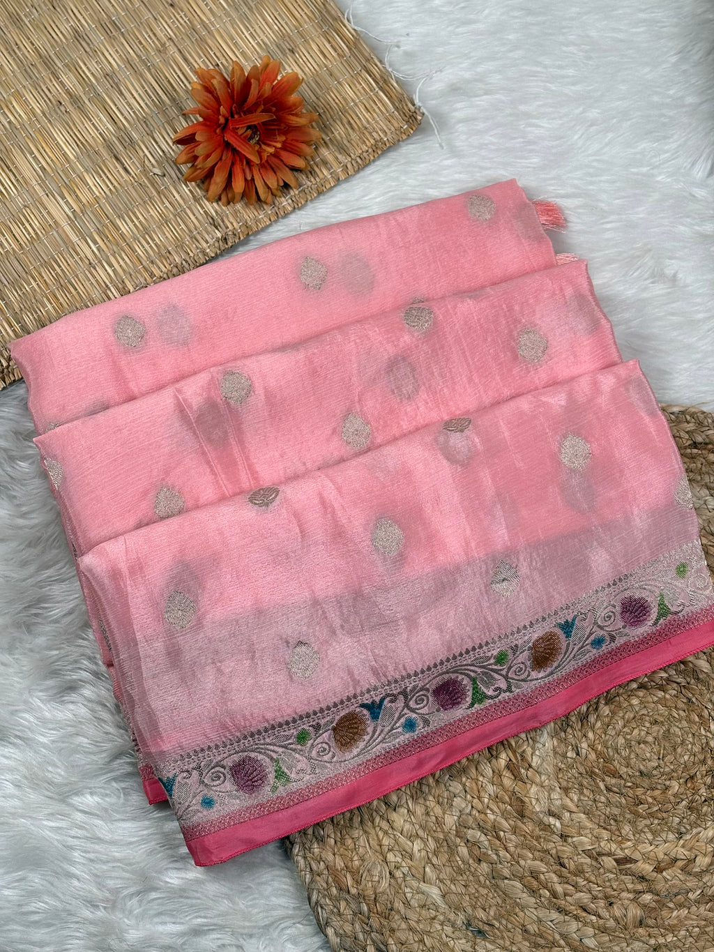 A folded pink saree with small metallic dot motifs and a decorative multicolor border, displayed on a white faux-fur surface with woven mat props.