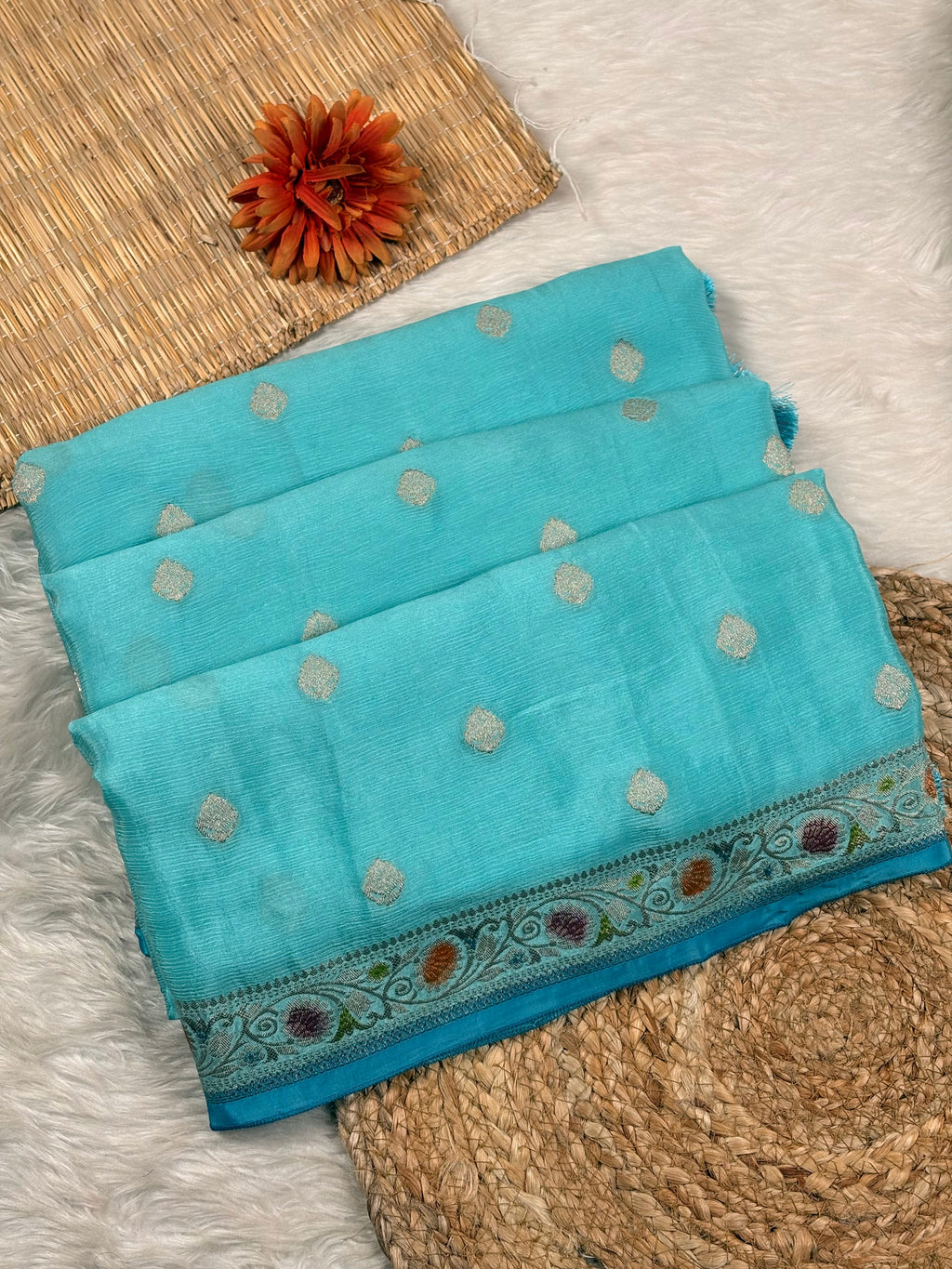 A turquoise blue folded saree with small metallic silver dots and a decorative woven border, styled on a white faux-fur surface with woven mats and an orange flower prop.