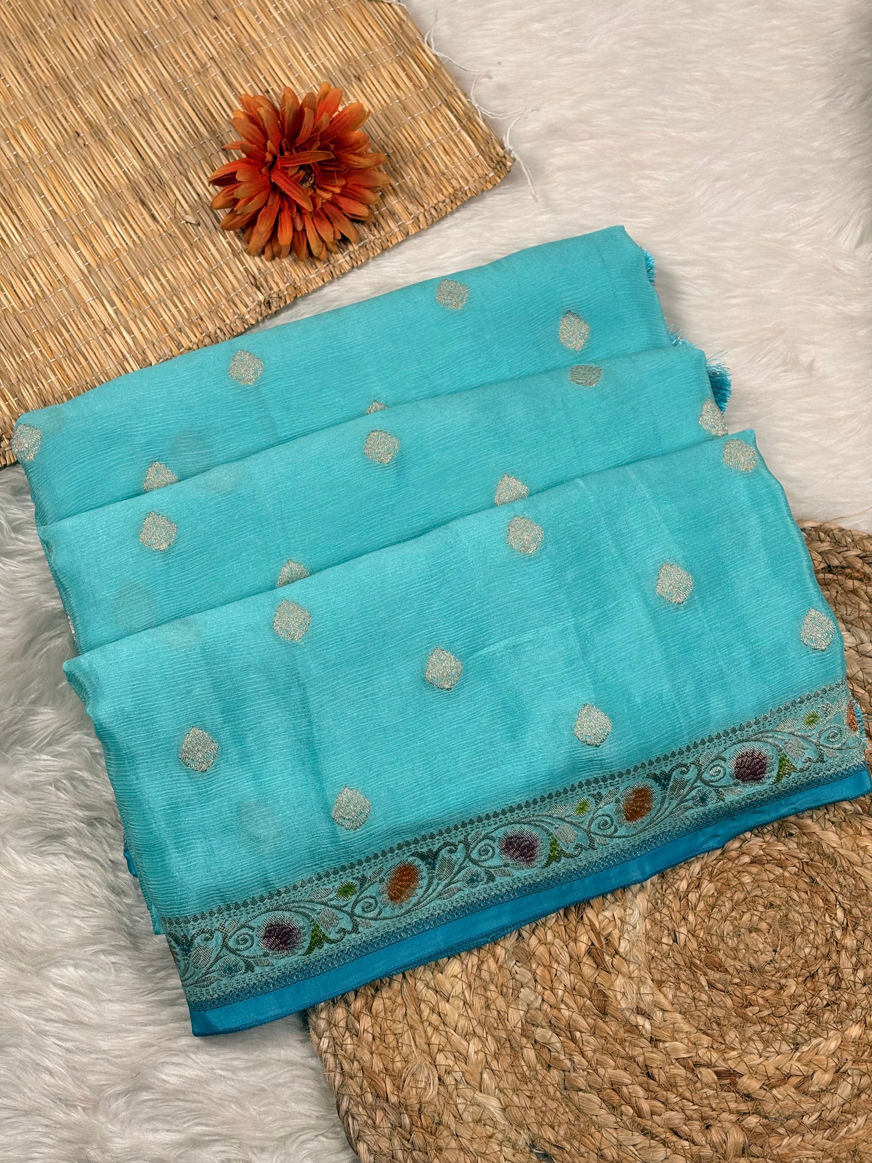 A turquoise blue folded saree with small metallic silver dots and a decorative woven border, styled on a white faux-fur surface with woven mats and an orange flower prop.