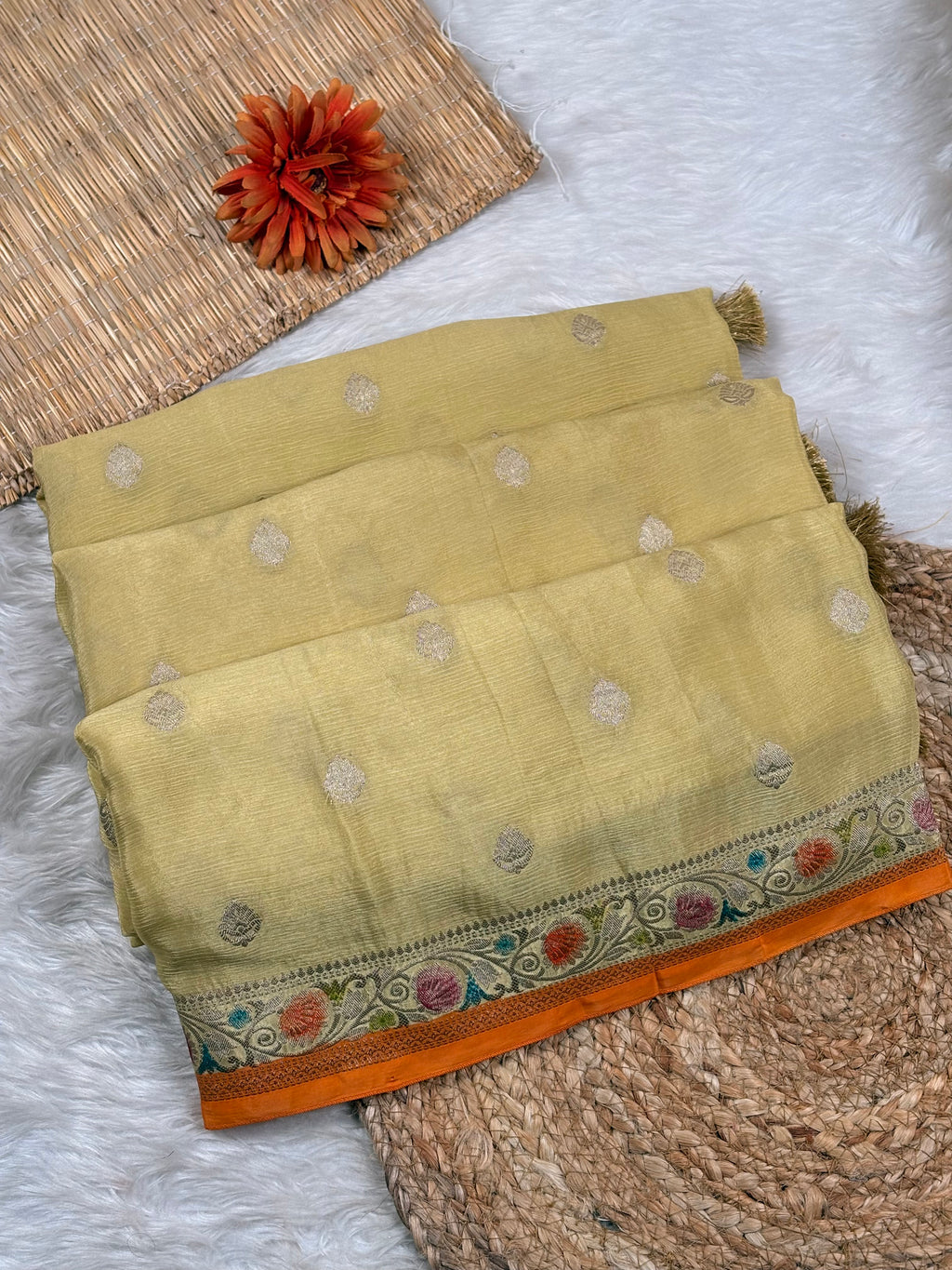 A folded olive green saree with silver  woven dots and a vibrant orange border, styled on a white faux fur rug with a woven mat and a decorative orange flower.