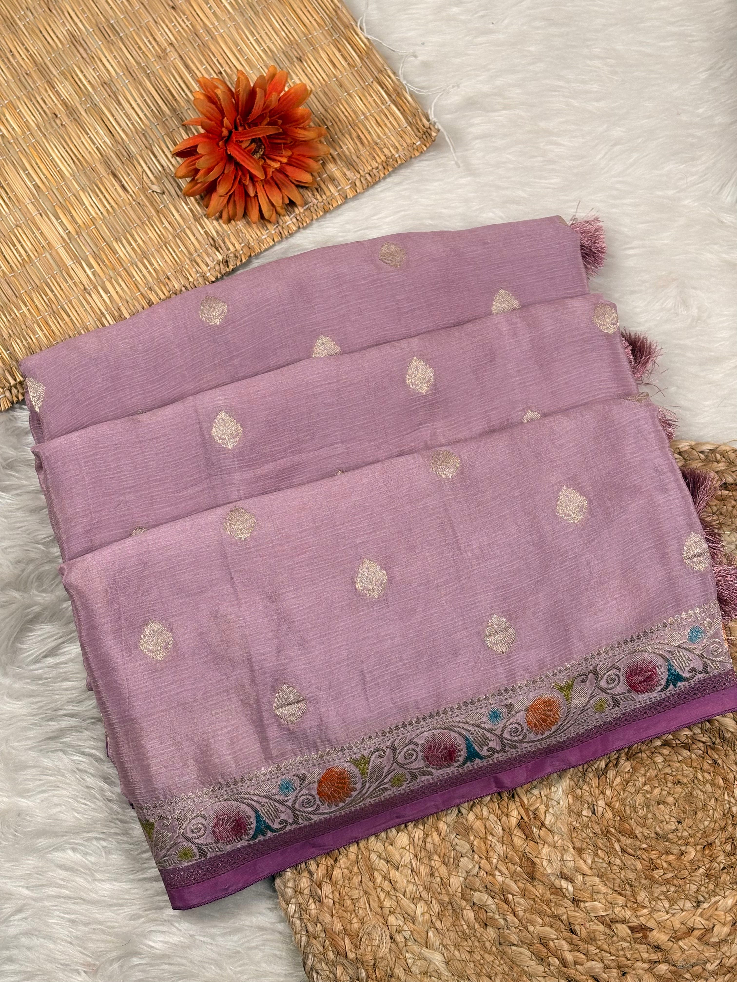 A folded lavender purple saree with small metallic gold dot motifs and a colorful woven border, displayed on a white faux fur surface with a woven mat and orange flower prop.