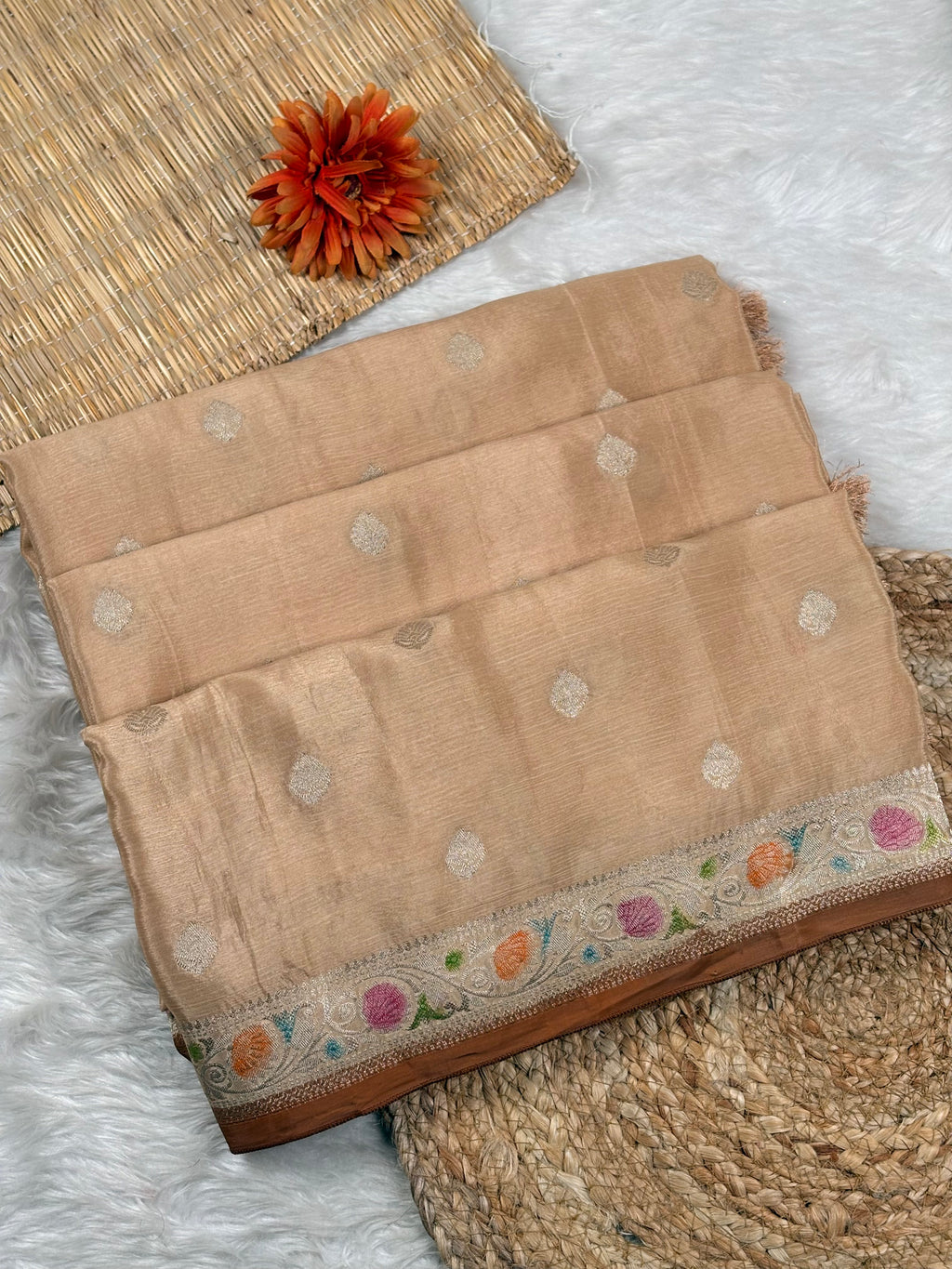 A folded beige silk saree with small metallic water gold dots and a colorful floral woven border, displayed on a white faux fur rug with a woven mat and a decorative orange flower.