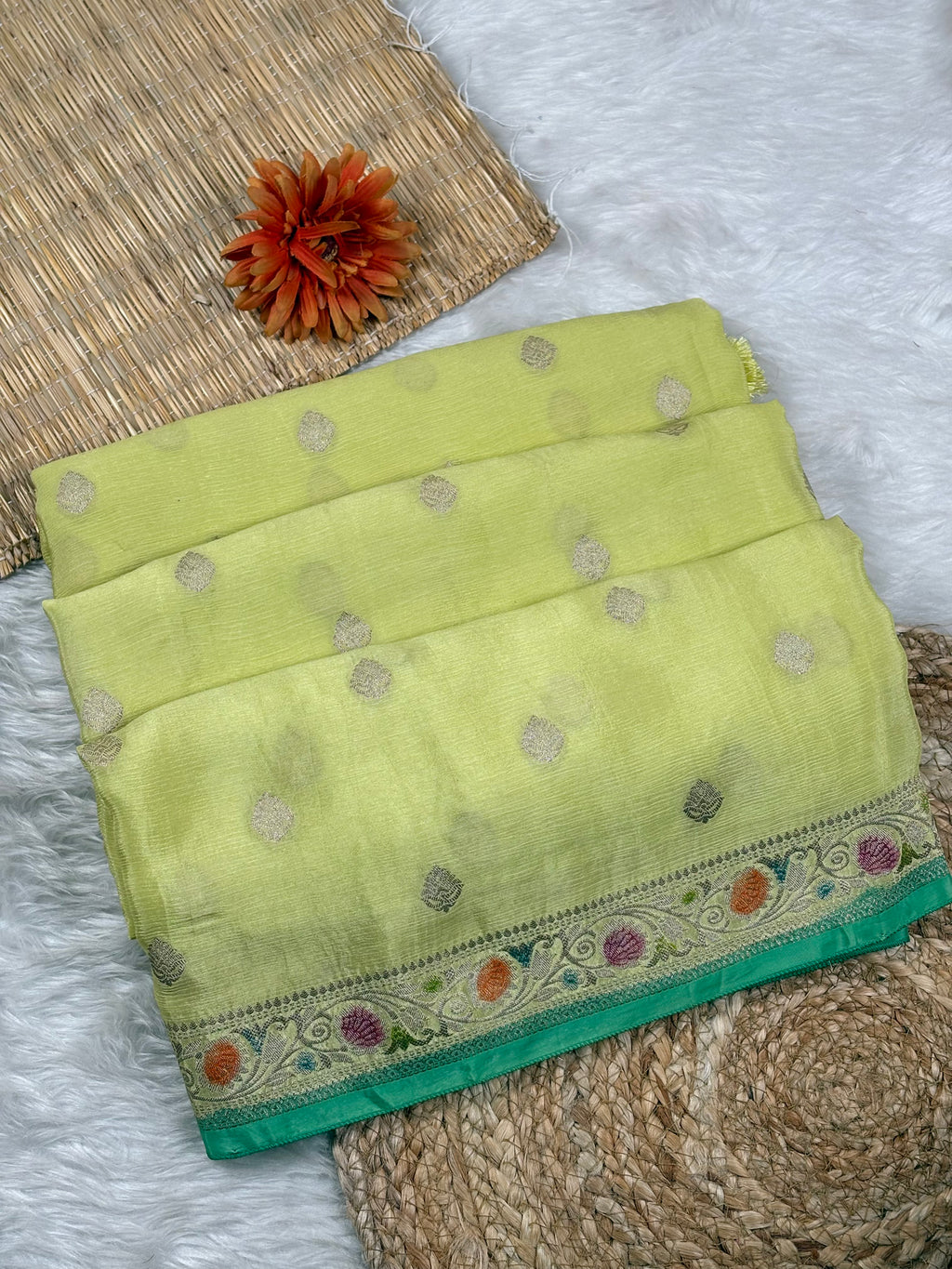 A folded lime green saree with small metallic gold dots and a decorative multicolor floral border, displayed on a white faux-fur surface with woven mat props and an orange flower accent.