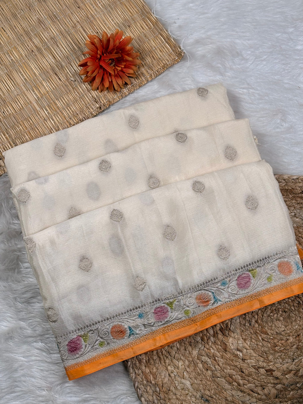 A folded off-white saree with small zari woven dots and a vibrant orange border featuring multicolored floral weaving, styled on a white faux fur surface with a woven mat and a decorative orange flower.