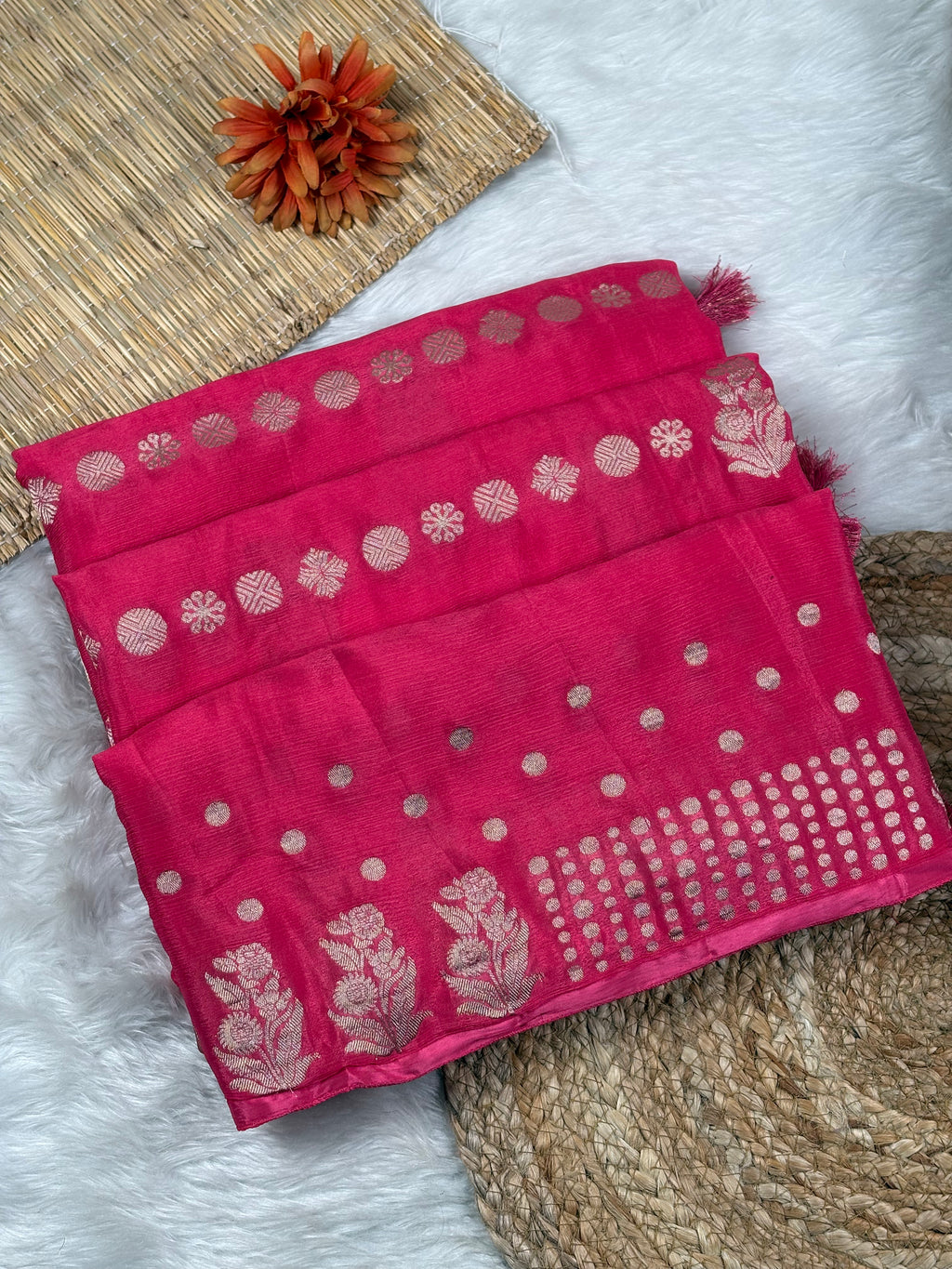 A folded bright pink saree with metallic gold floral and circular motifs, displayed on a white faux-fur surface with woven mat props.