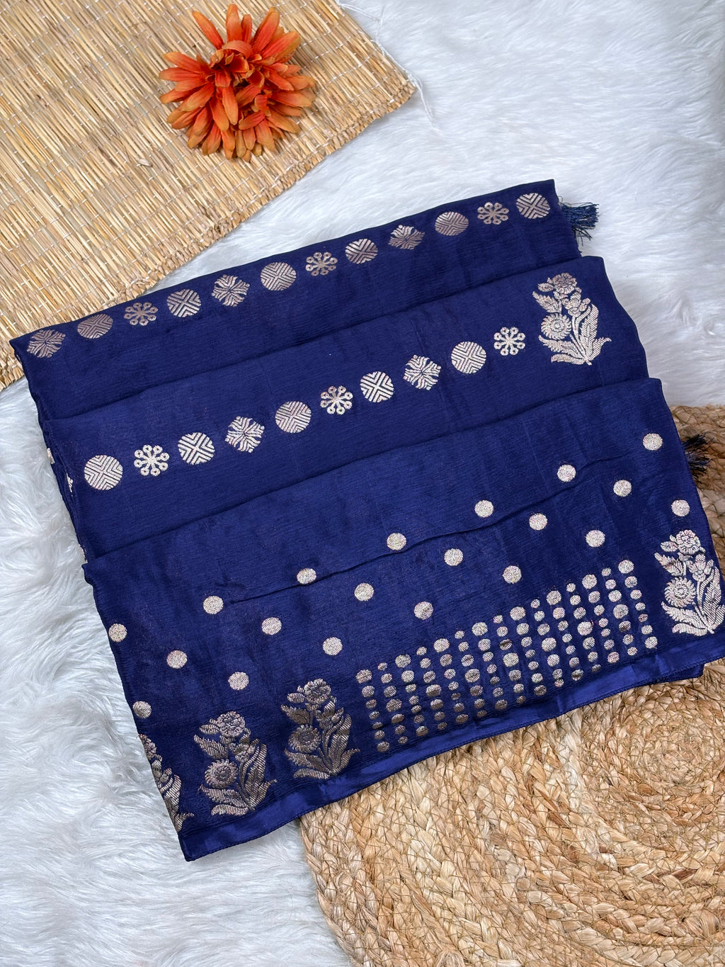 A folded navy blue saree, decorated with water gold metallic floral and geometric weaving, displayed on a white faux fur surface with a woven mat and orange flower prop.