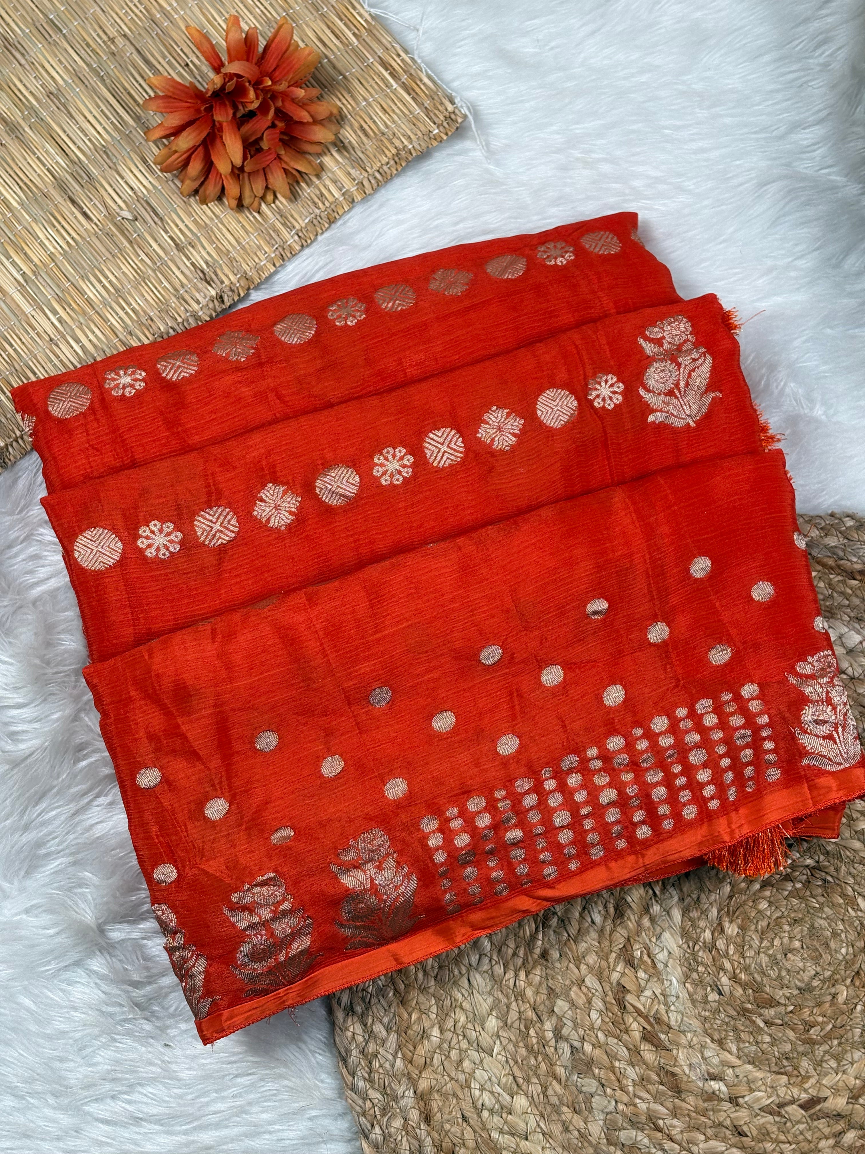 A folded red saree with water gold zari floral and geometric motifs, displayed on a white faux-fur surface with woven mats and a dried orange flower as styling props.