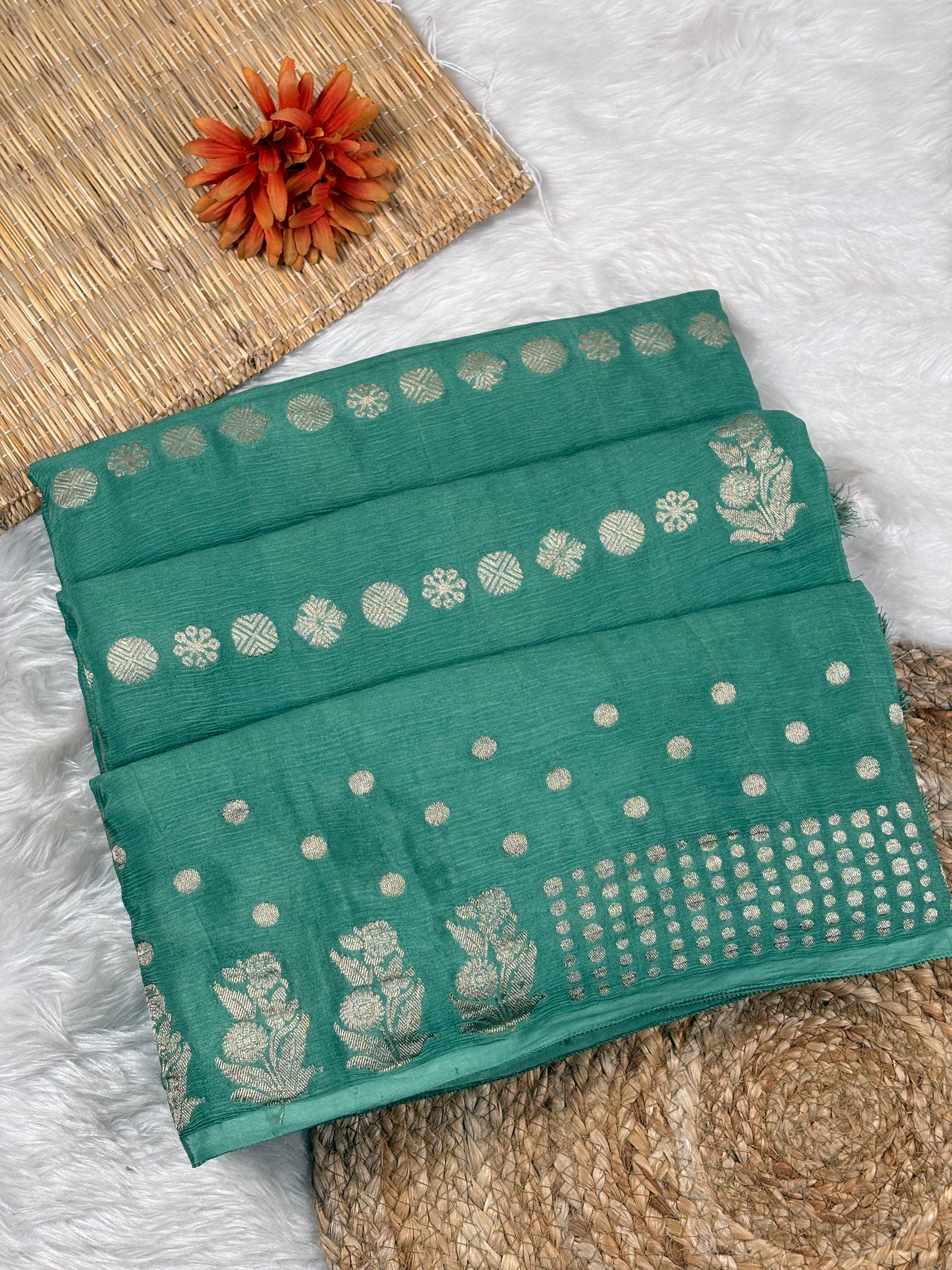 A folded teal green saree with metallic water gold floral and dot motifs, displayed on a white faux fur surface with a woven mat and orange flower prop.