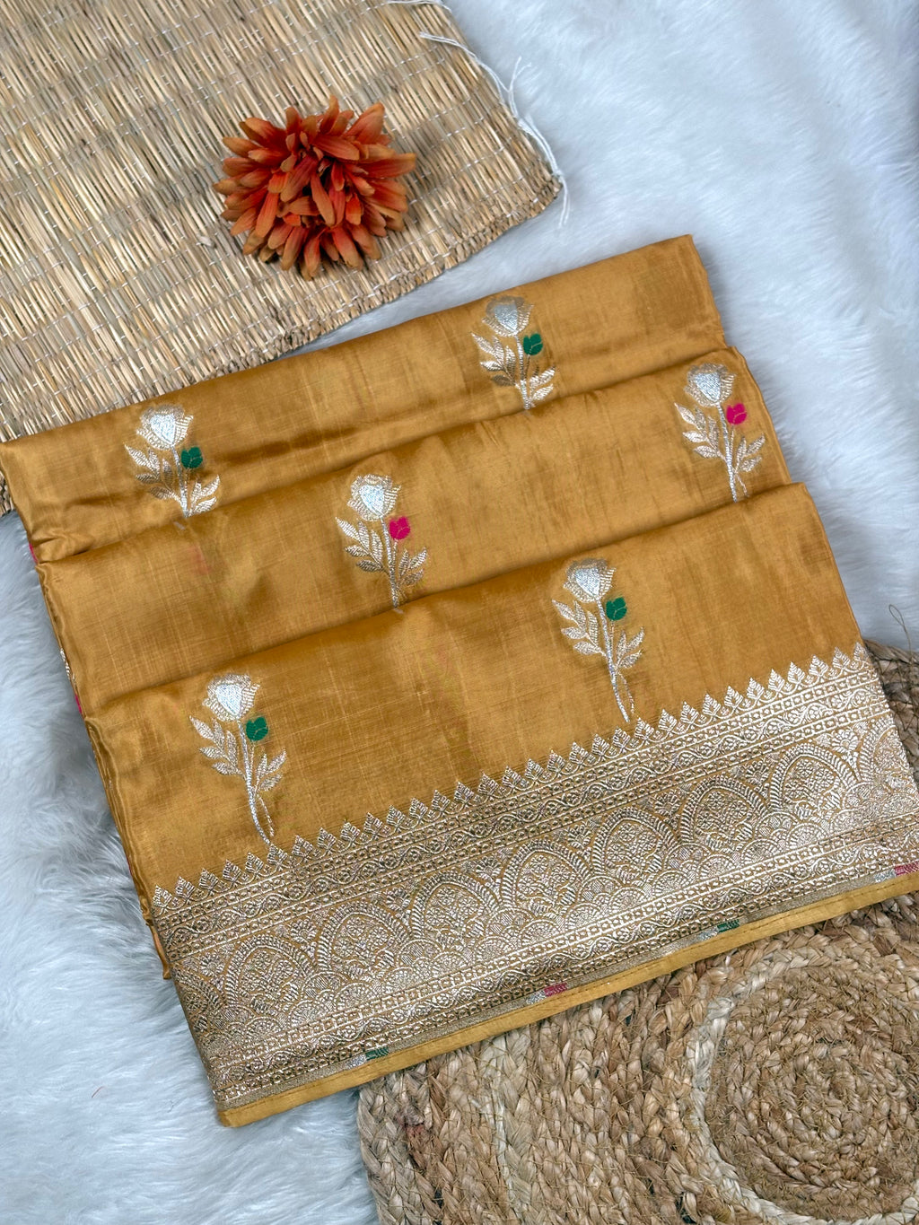 A folded mustard yellow silk saree with a wide gold zari border and woven floral motifs, displayed on a white faux fur surface with decorative props.