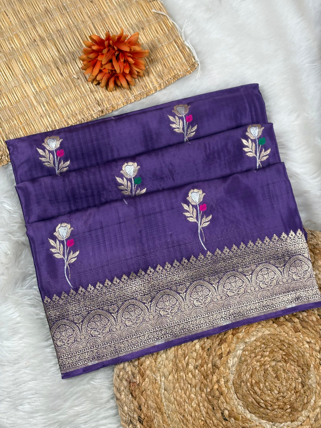 A folded purple silk saree with silver zari floral weaving and a wide decorative border, styled on a white faux fur surface with woven mats and an orange flower prop.