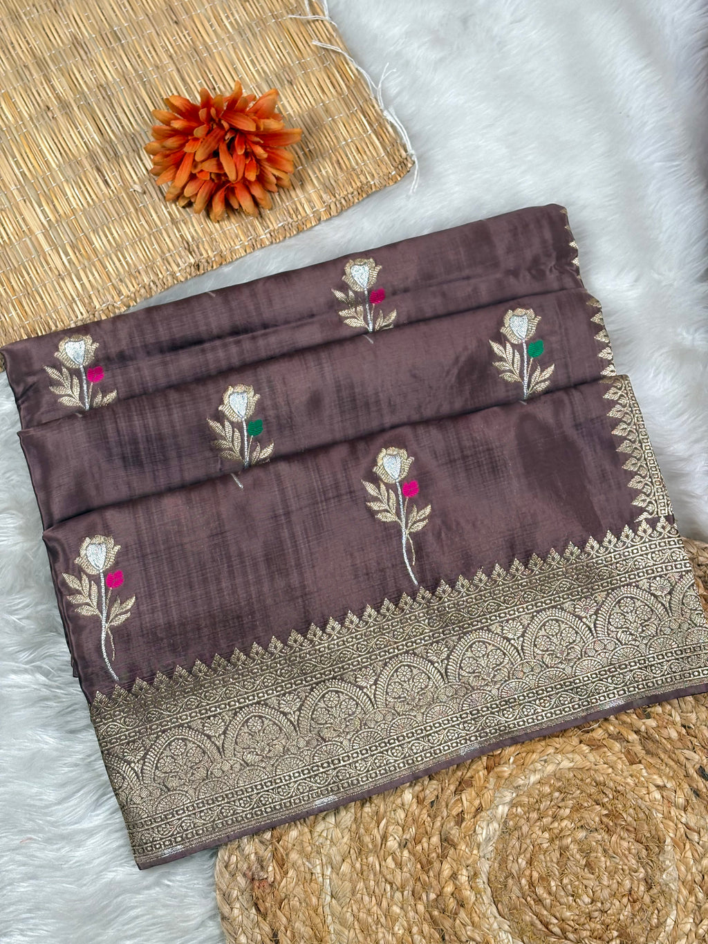 A folded dark brown silk saree with intricate floral weaving and a wide metallic gold border, styled on a white faux fur surface with woven mats and an orange flower.