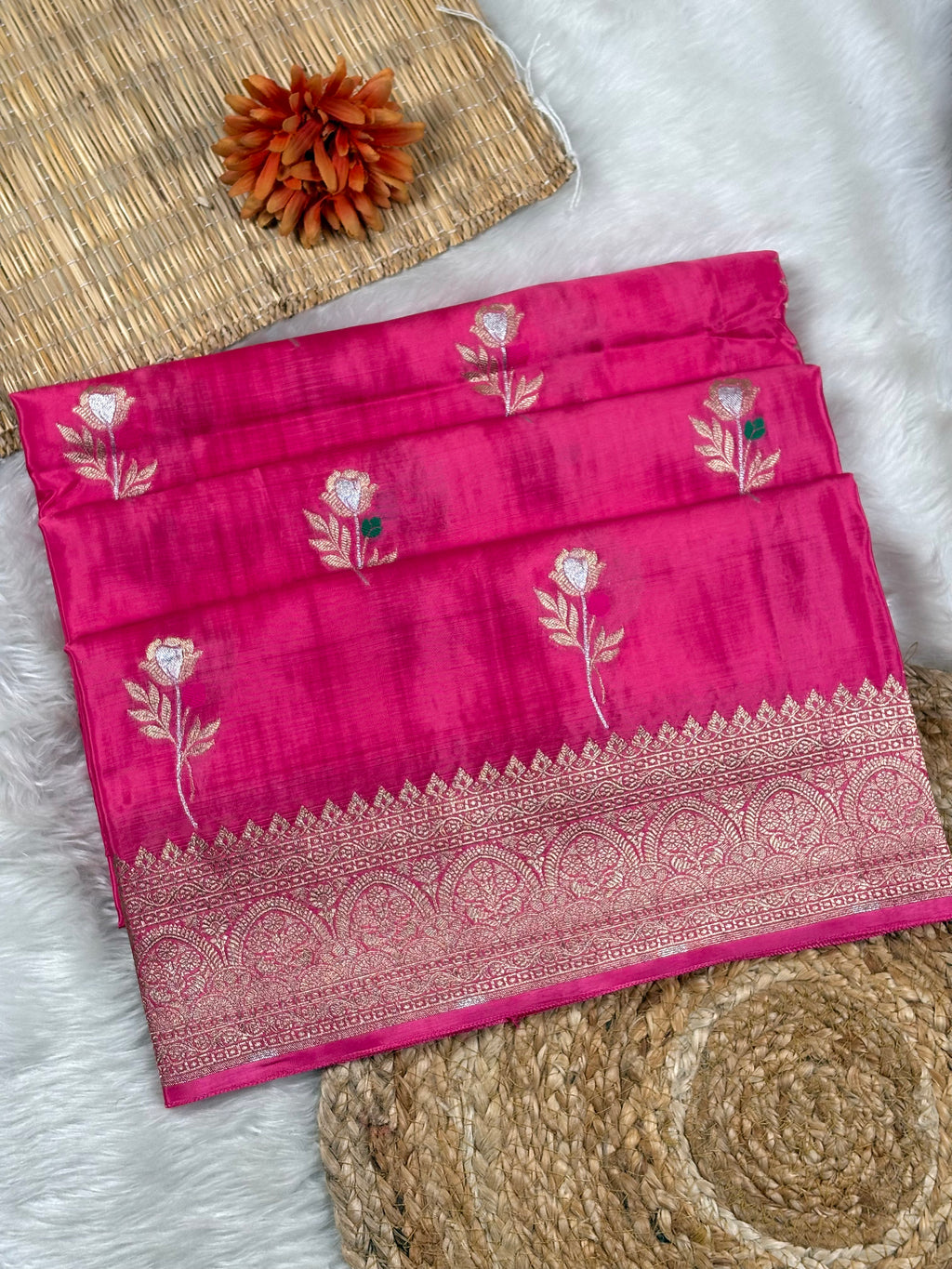 A folded bright pink silk saree with floral weaving and a detailed gold zari border, displayed on a white faux fur surface with woven mat props.
