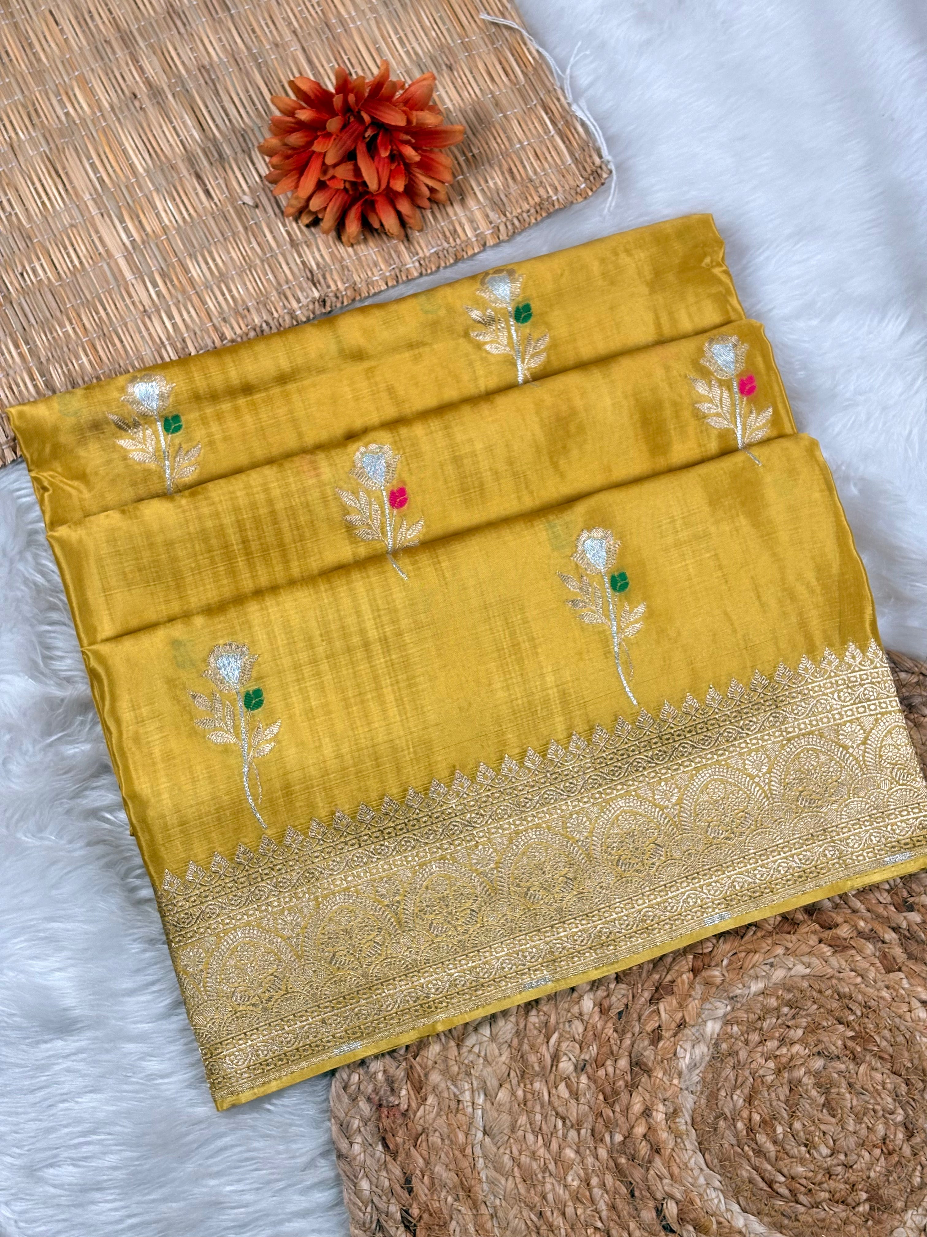 A folded mustard yellow silk saree with intricate gold zari waving and small floral motifs, displayed on a white faux fur surface with decorative woven mats.