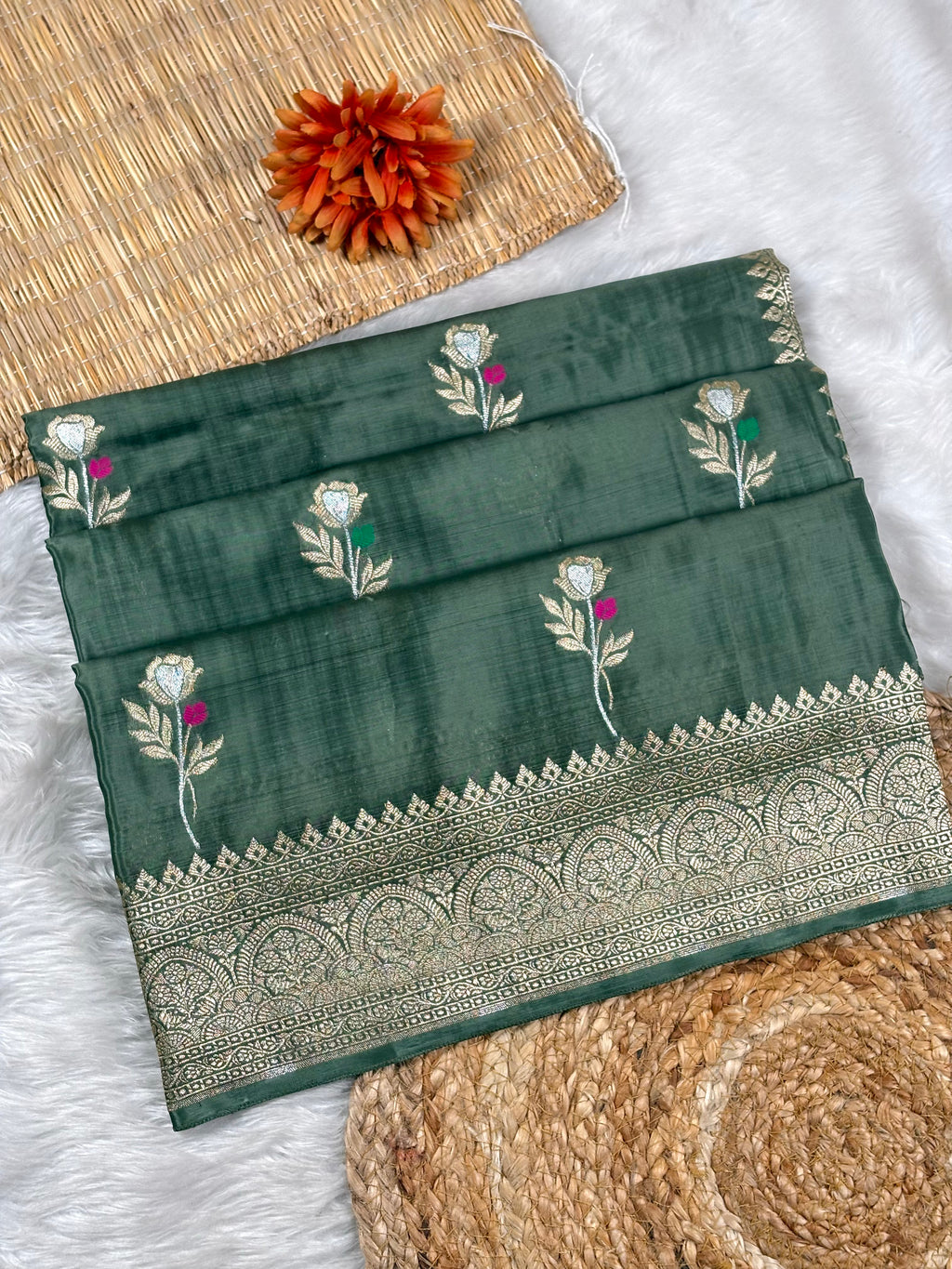 A folded dark green saree with floral weaving and a wide ornate metallic border, styled on a white faux-fur surface with woven mats and an orange flower prop.