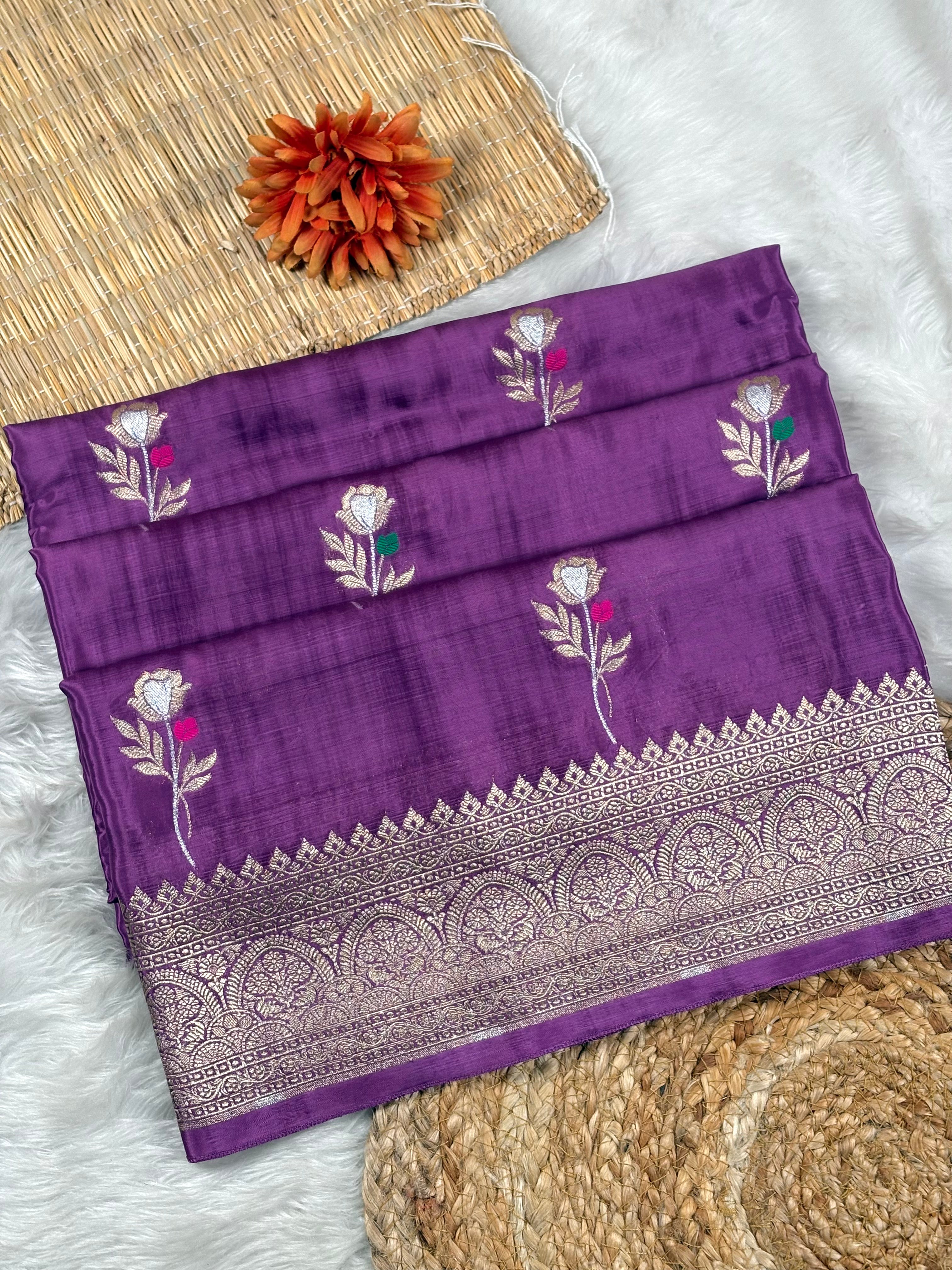 A folded purple silk saree with floral weaving and a wide metallic border, displayed on a white faux fur surface with decorative woven mats and an orange flower.