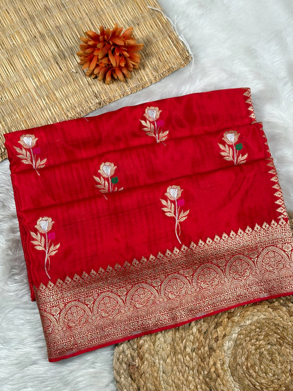 A folded red silk saree with intricate gold zari woven and floral motifs, displayed on a white faux fur surface with a woven mat and decorative flower in the background.