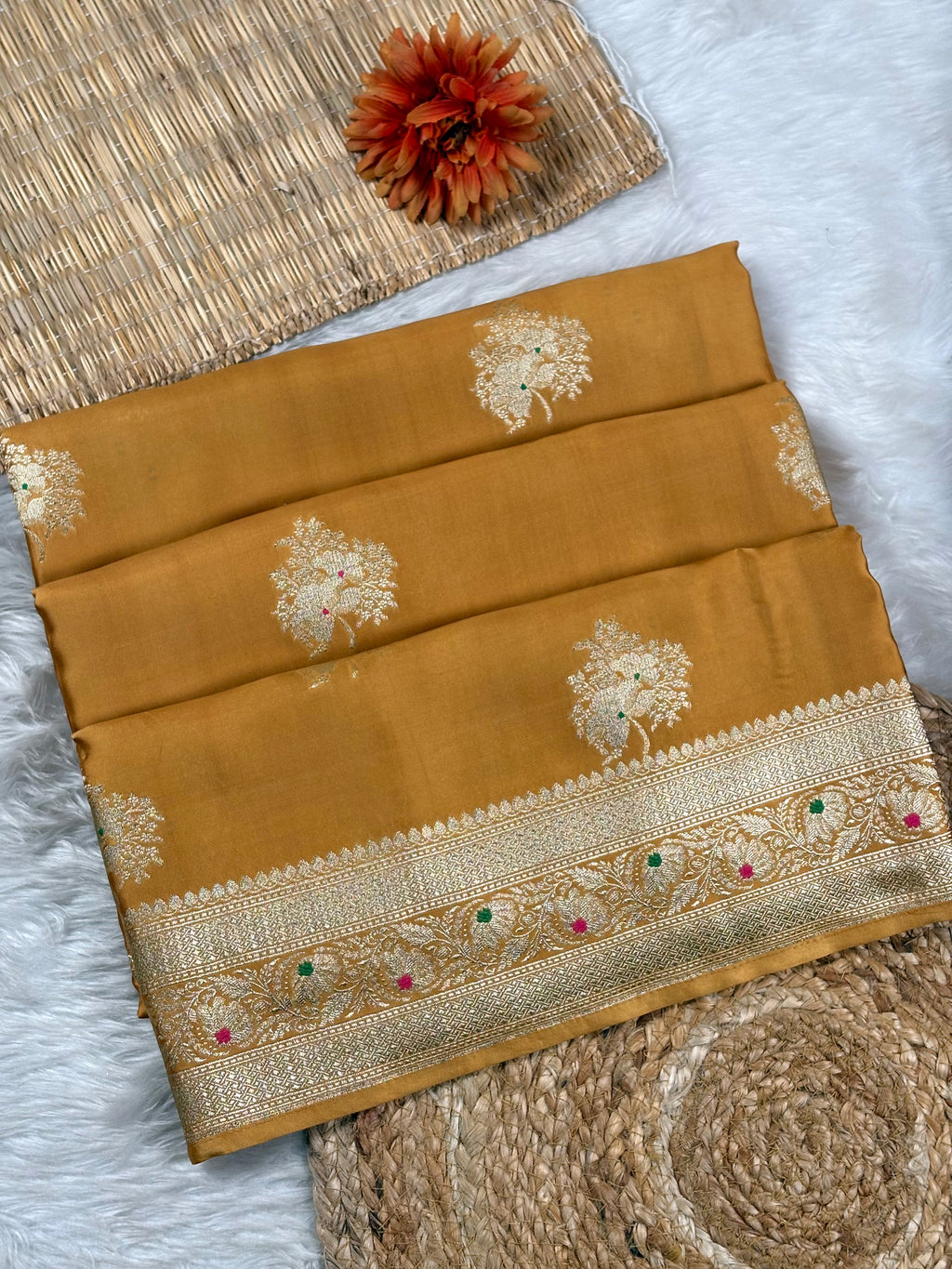 A folded mustard yellow saree with gold floral motifs and a wide ornate gold zari border decorated with small green and pink mina accents, styled on a white faux fur surface with woven mats and a dried orange flower.