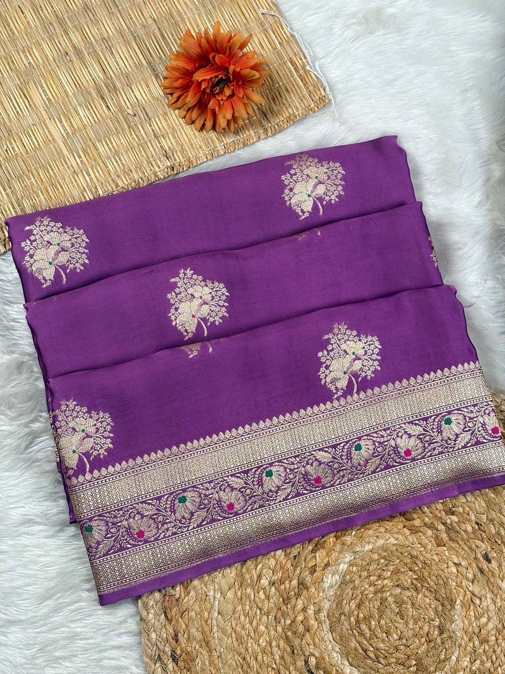 A folded purple silk saree with intricate gold floral motifs and a decorative border, displayed on a white faux fur surface with a woven mat and orange flower as props.