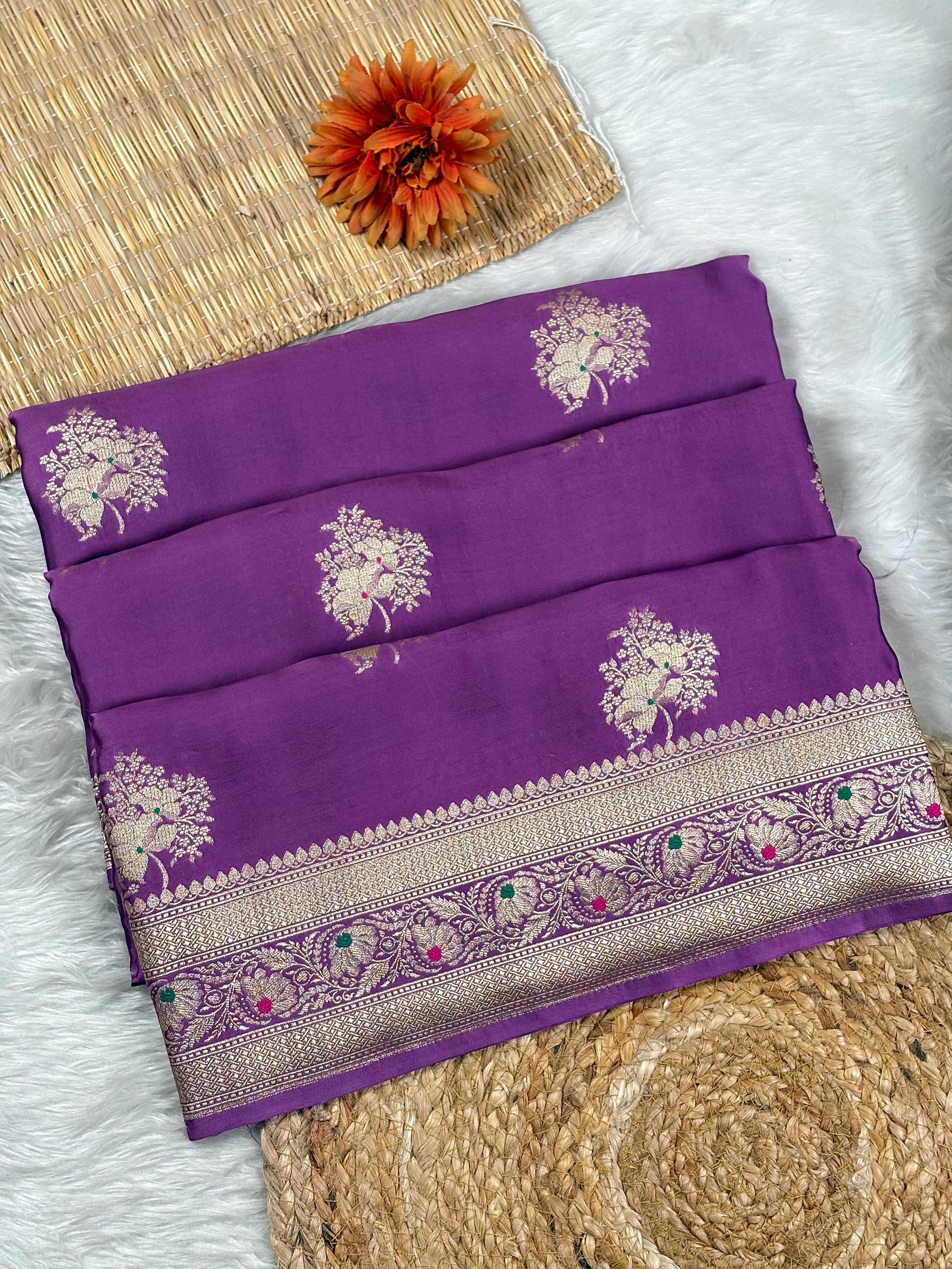 A folded purple silk saree with intricate gold floral motifs and a decorative border, displayed on a white faux fur surface with a woven mat and orange flower as props.
