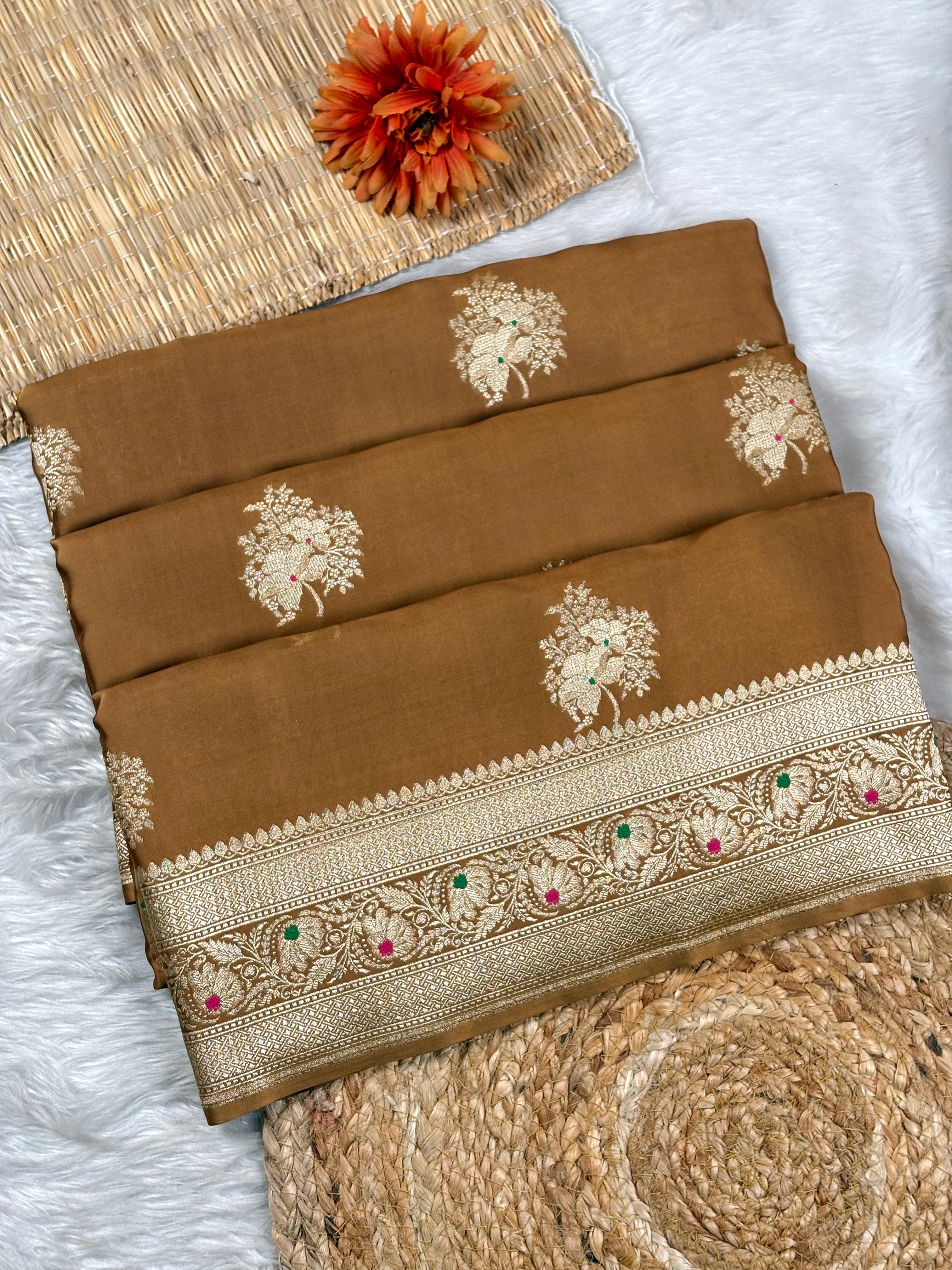 A folded brown saree with gold floral weaving and a decorative border featuring small pink and green accents, styled on a white faux-fur surface with woven mats and an orange flower.