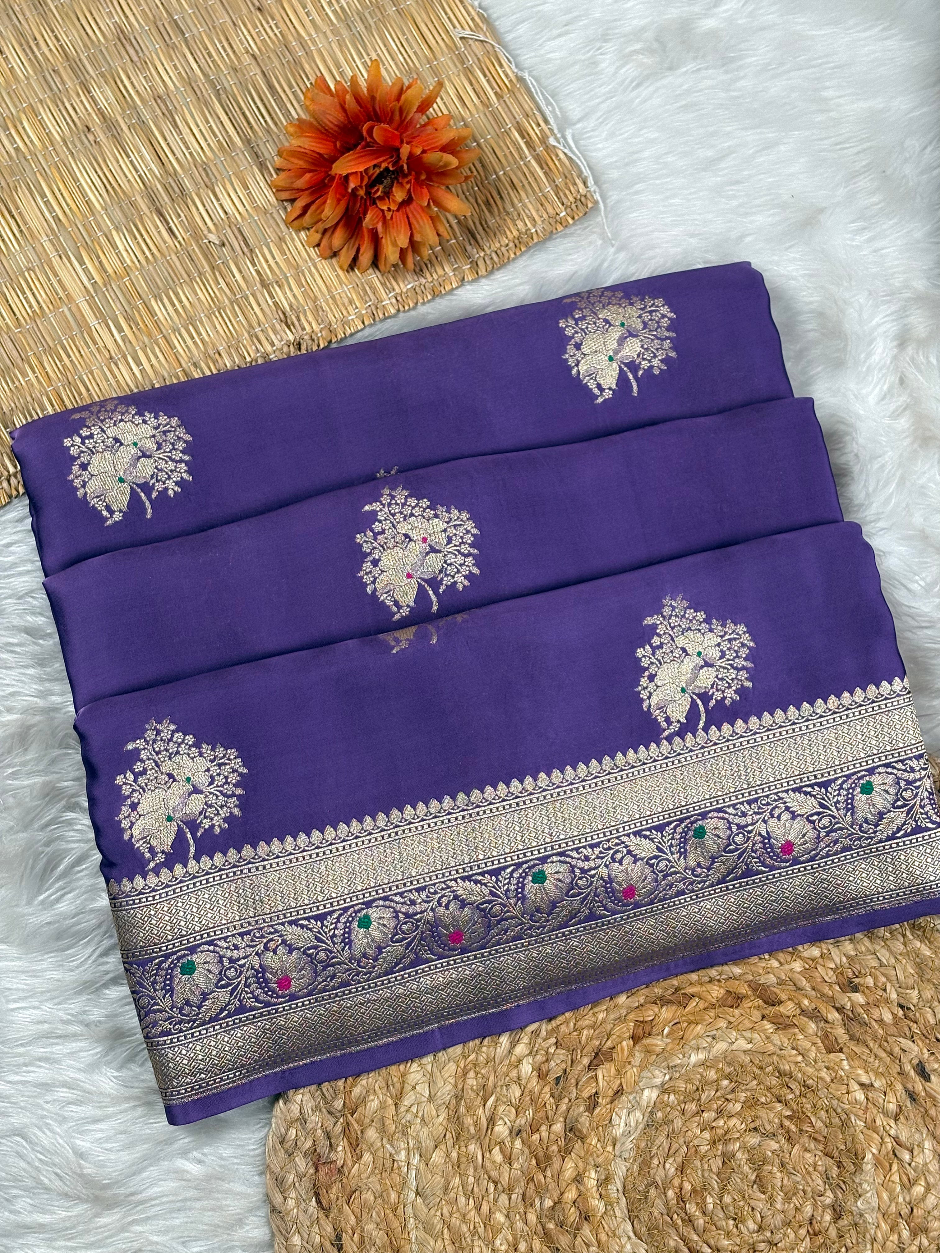 A folded purple silk saree with gold zari weaving, featuring floral motifs and a detailed border, displayed on a white faux fur surface with a woven mat and an orange flower as props.