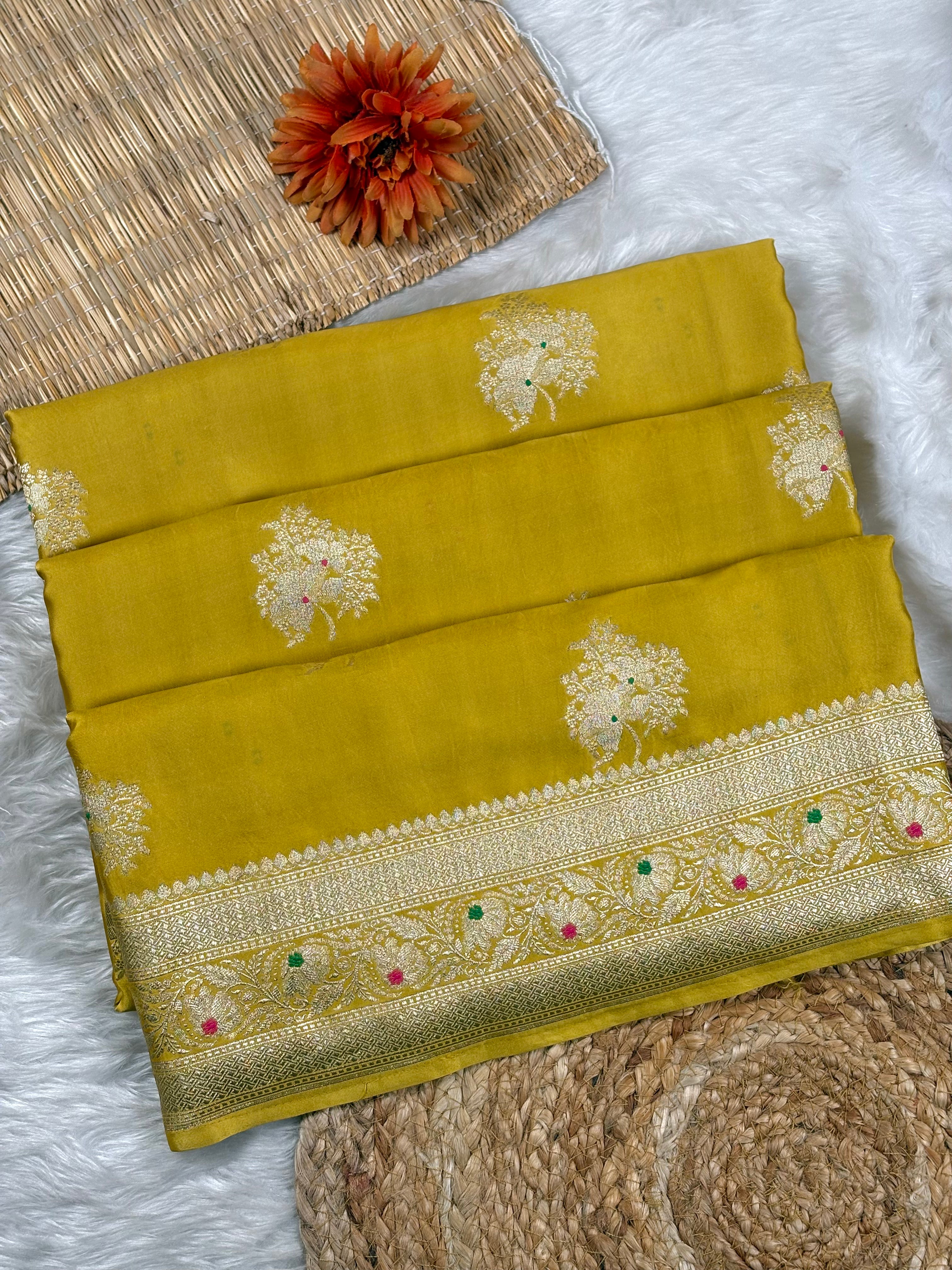 A folded olive yellow saree with gold floral weaving and a decorative gold zari border, styled on a white faux fur surface with a woven mat and a dried orange flower.