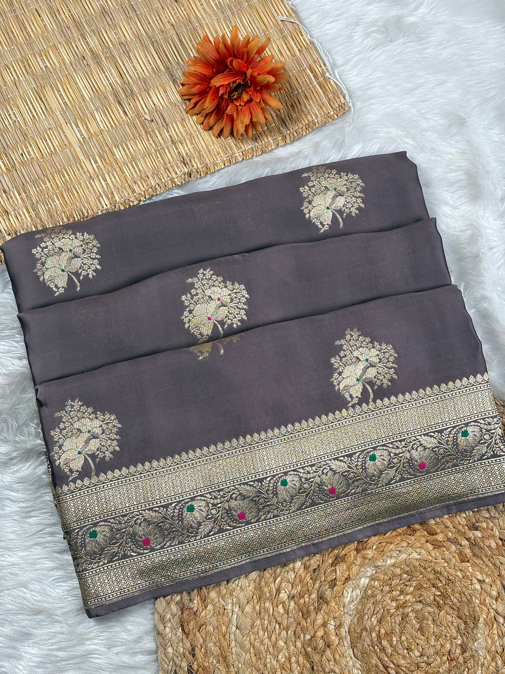 A folded dark gray saree with gold floral motifs and a decorative gold-toned border, styled on a white faux-fur surface with woven mats and an orange flower prop.