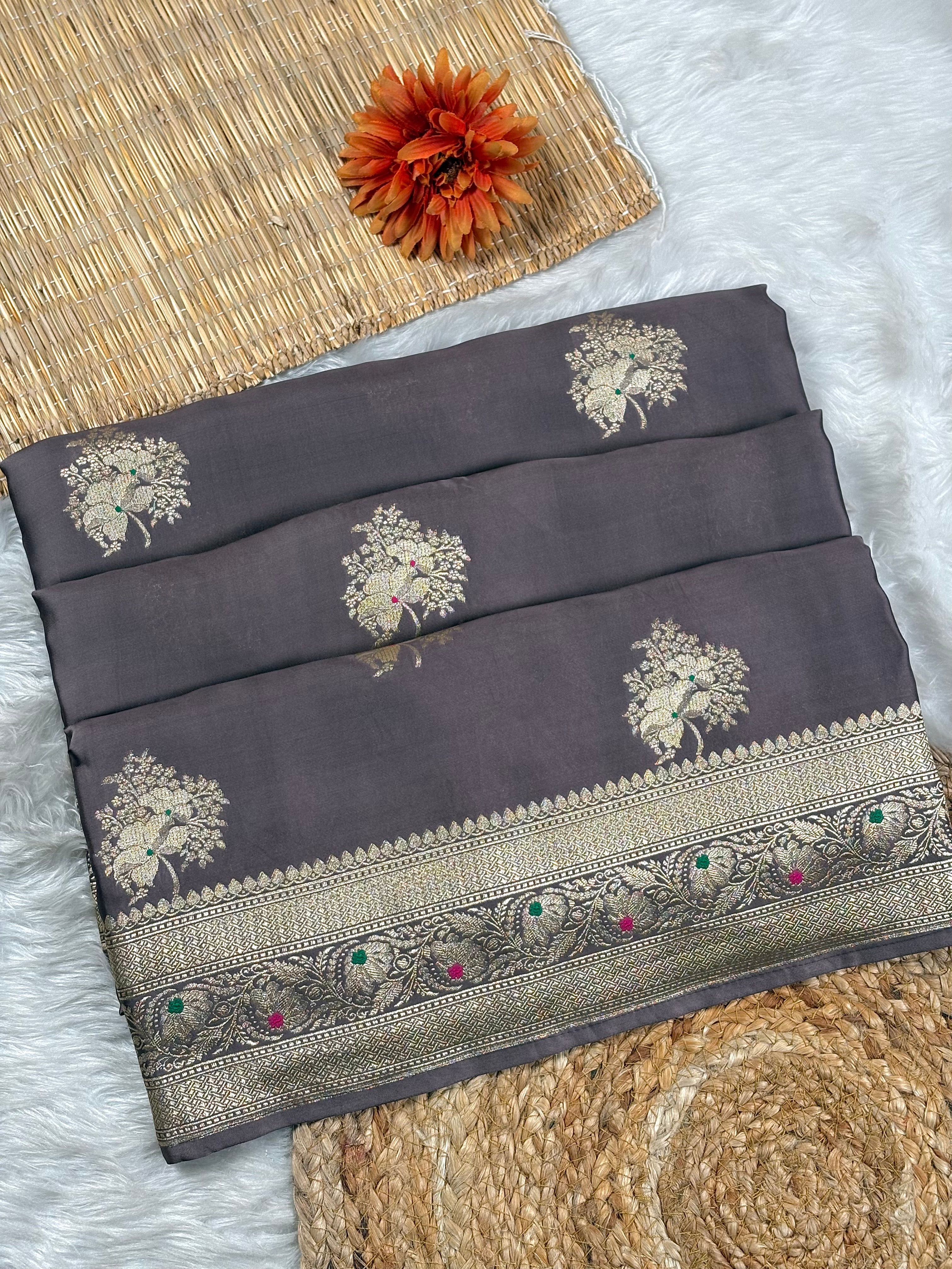A folded dark gray saree with gold floral motifs and a decorative gold-toned border, styled on a white faux-fur surface with woven mats and an orange flower prop.