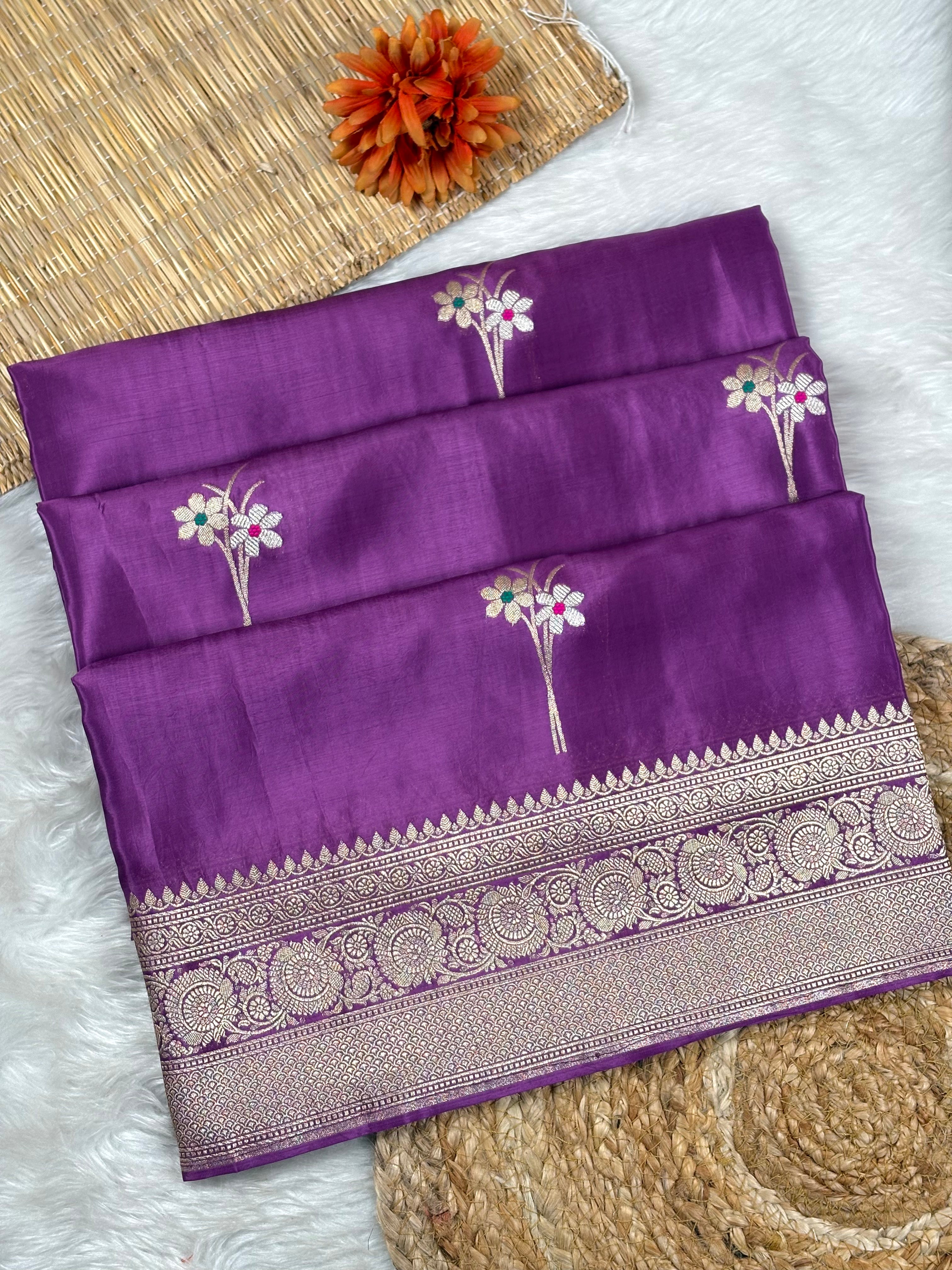 A folded purple silk saree with a wide silver zari border and floral motifs, displayed on a white faux fur surface with a woven mat and orange flower prop.