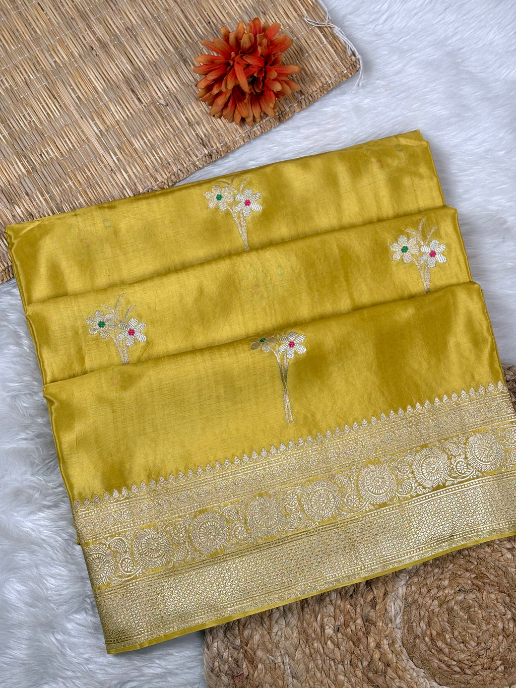 A folded yellow silk saree with a wide gold zari border and small tilfi mina woven floral motifs, displayed on a white faux-fur surface with woven props.
