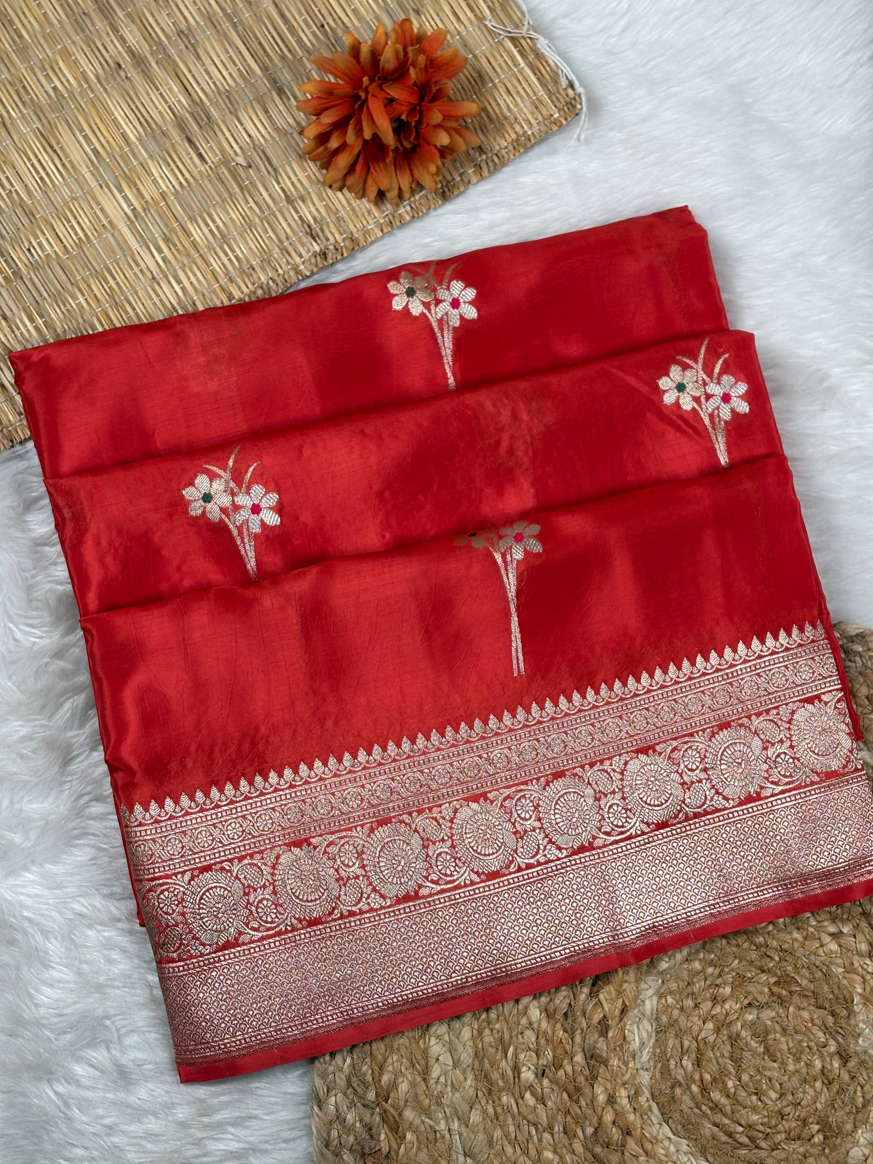 A folded red silk saree with tilfi mina floral weaving and a detailed gold border, displayed on a white faux fur surface with a woven mat and a dried orange flower as props.
