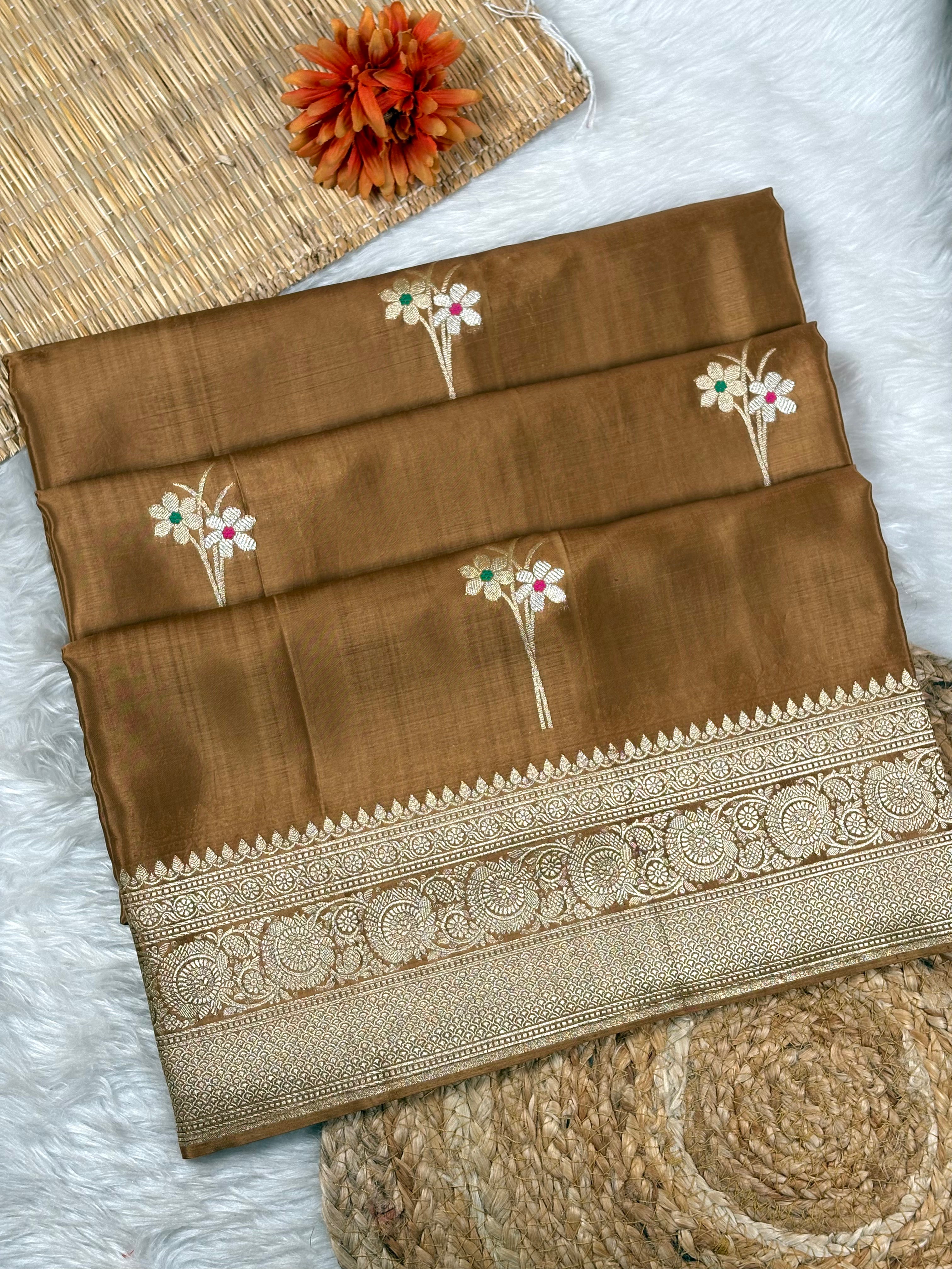A folded brown saree with a wide ornate gold border and small woven floral motifs, displayed on a white faux-fur surface with woven props and a decorative orange flower.