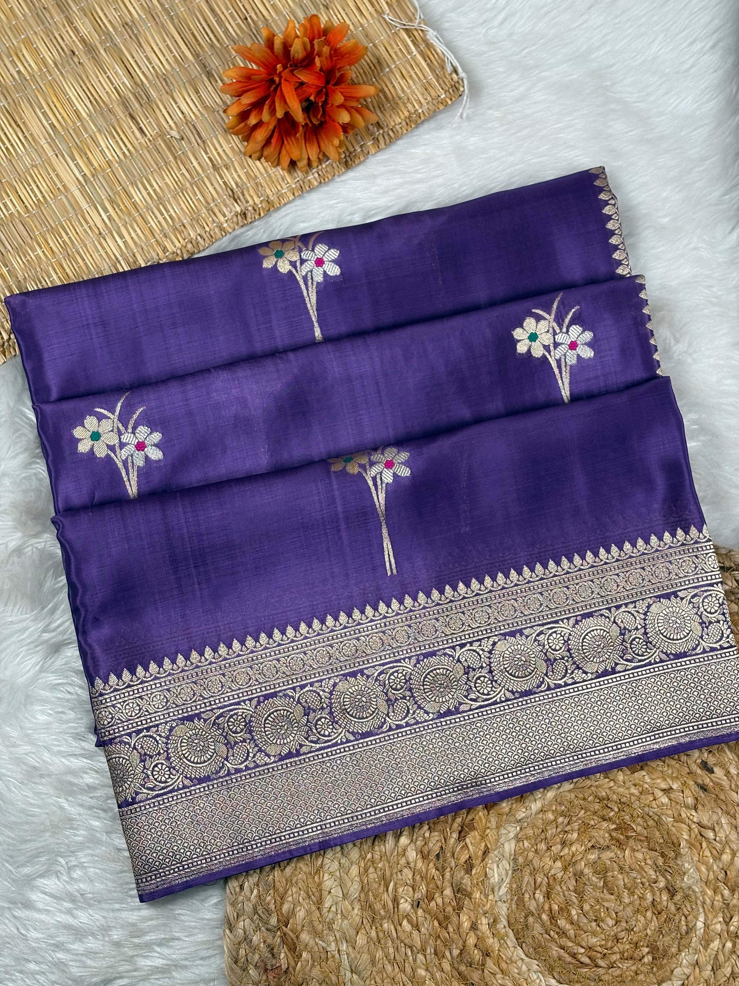 A folded purple silk saree with floral zari weaving, displayed on a white faux fur surface with a woven mat and orange flower as props.