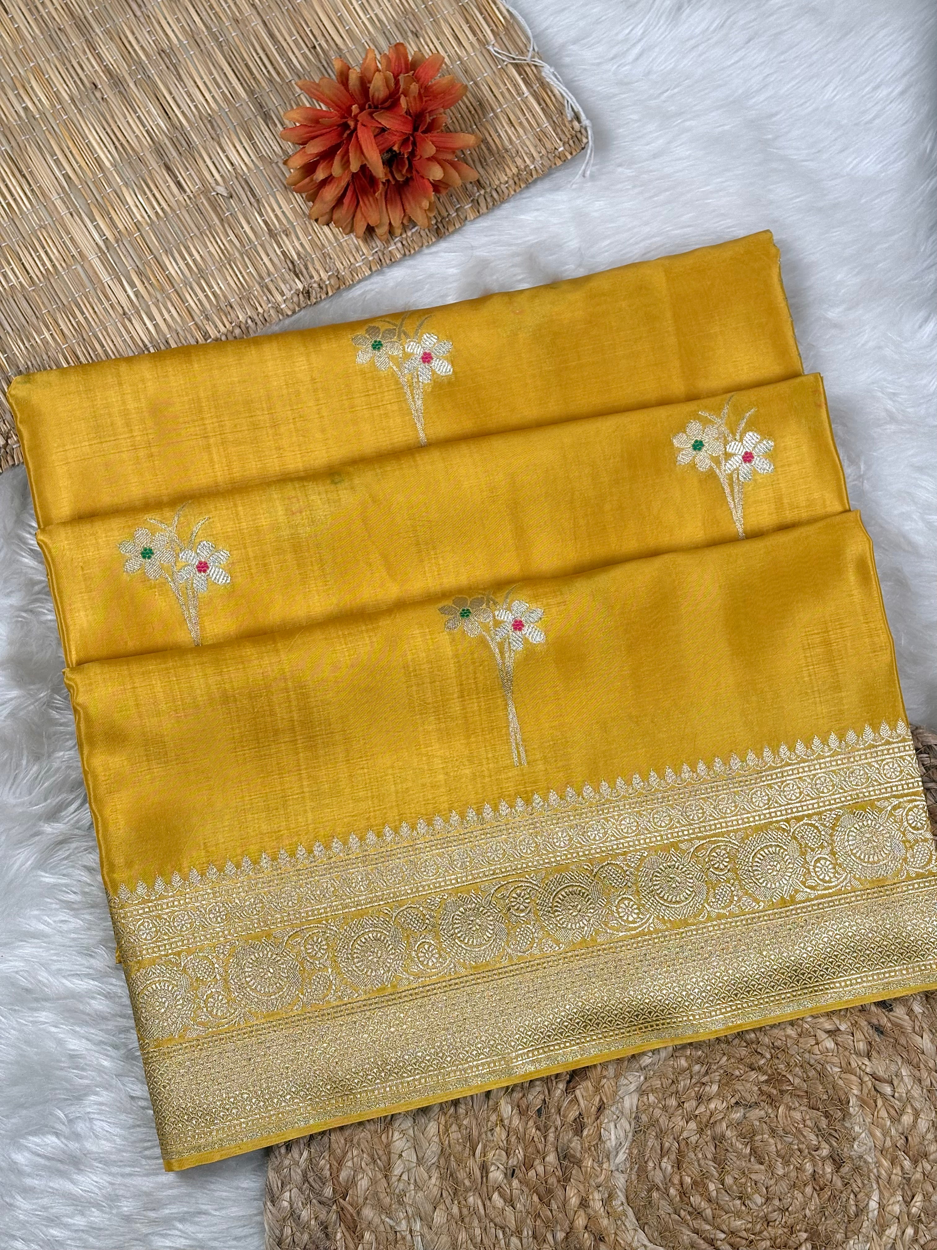 A folded mustard yellow silk saree with delicate floral weaving and a wide gold zari border, styled on a white faux fur surface with a woven mat and a small orange flower.