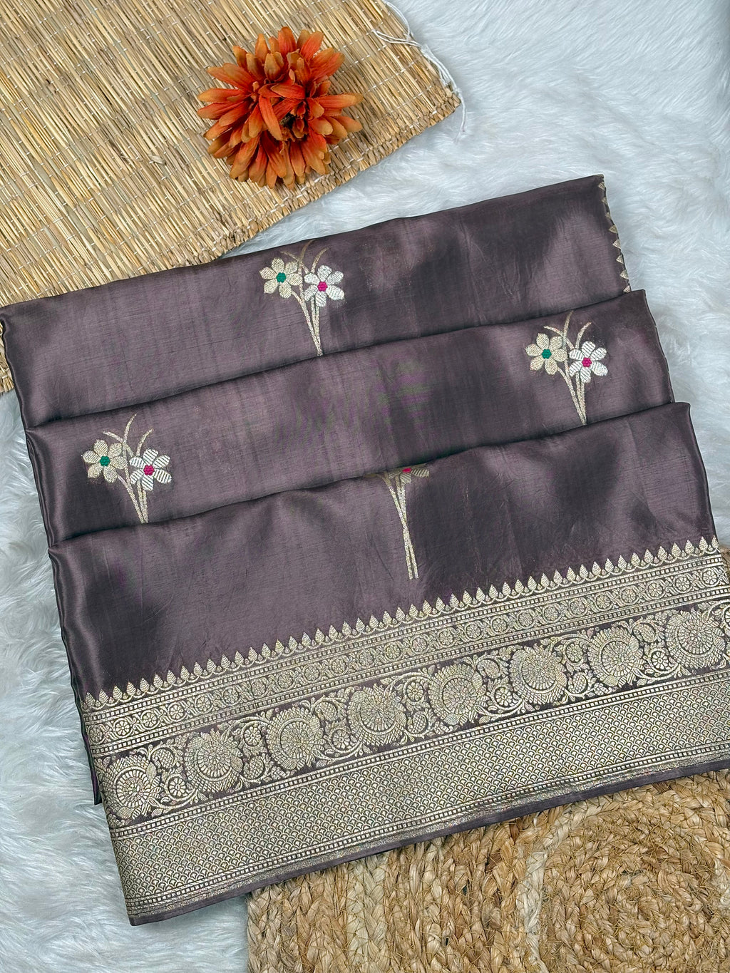 A folded dark plum purple silk saree with intricate gold zari border and floral motifs, displayed on a white faux fur surface with a woven mat and orange flower prop.