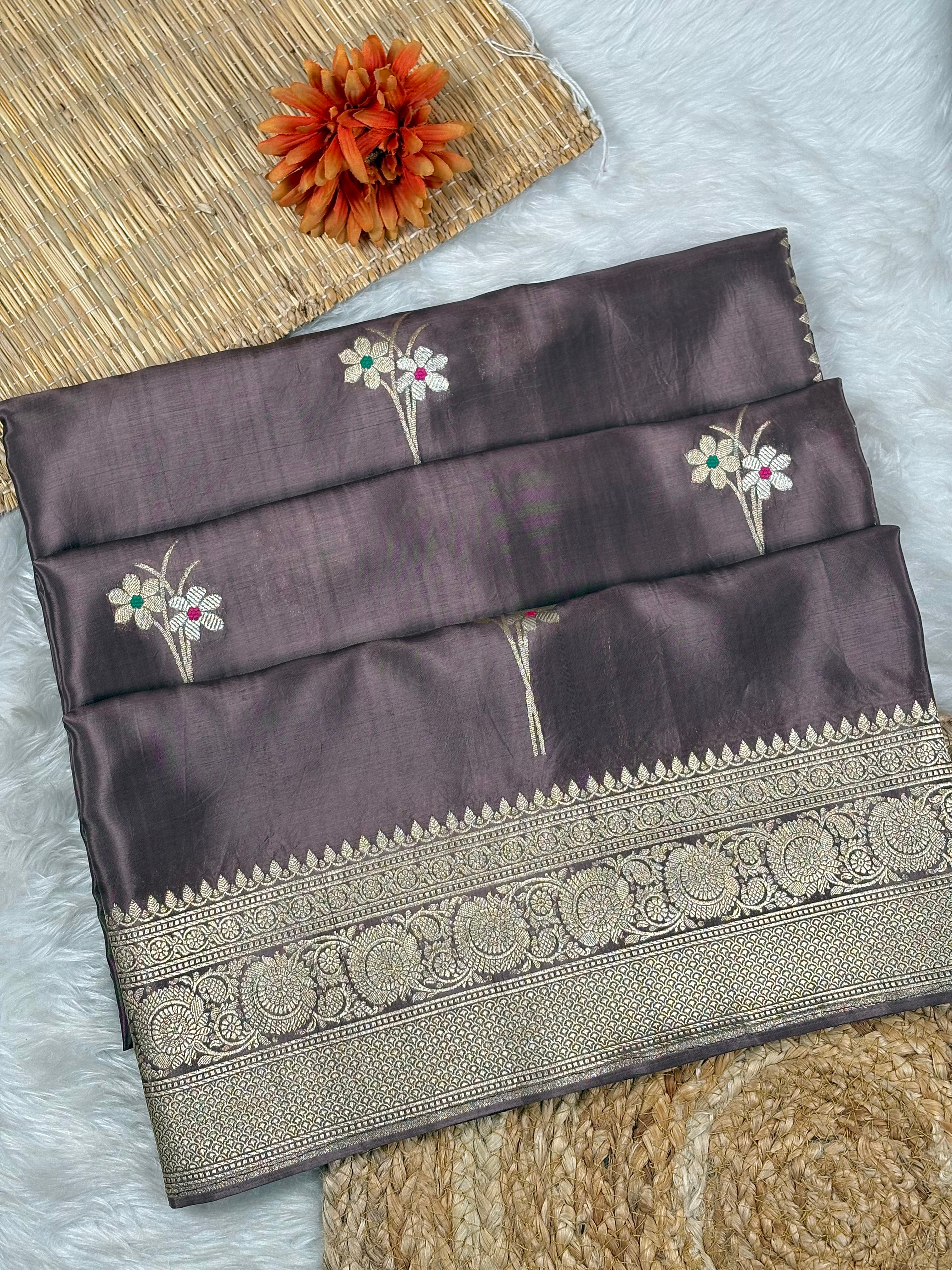 A folded dark plum purple silk saree with intricate gold zari border and floral motifs, displayed on a white faux fur surface with a woven mat and orange flower prop.