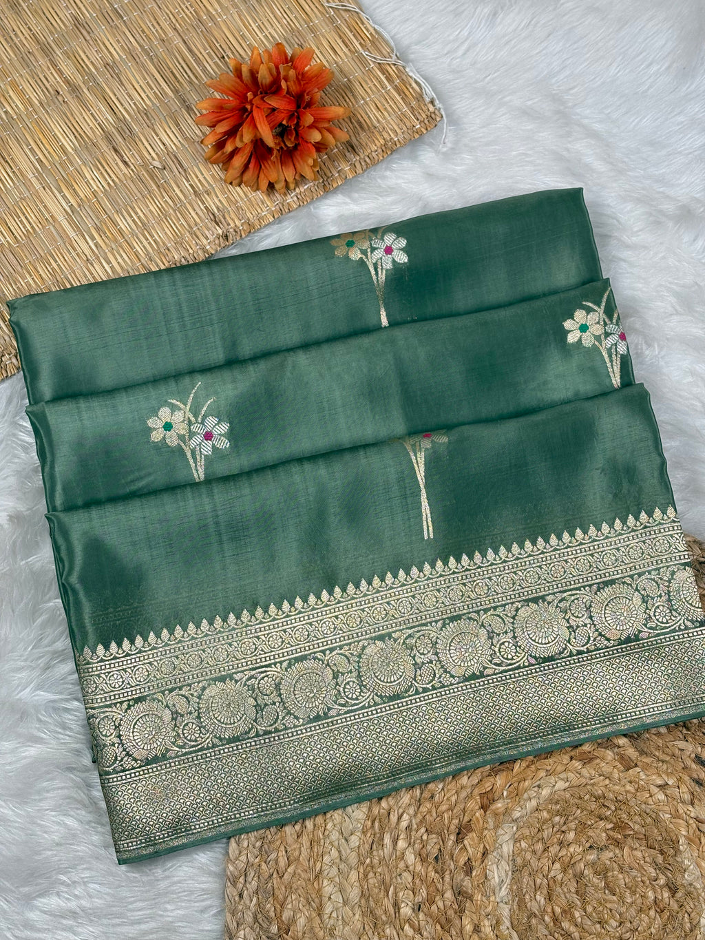 A folded dark green silk saree with intricate silver zari weaving and a wide decorative border, displayed on a white faux fur surface with a woven mat and orange flower as props.