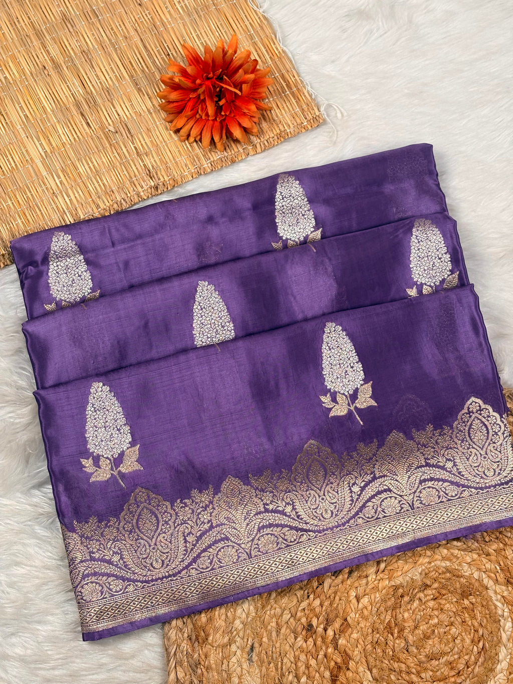 A folded purple silk saree with alfi mina floral woven motifs and a detailed gold zari border, displayed on a white faux fur surface with a woven mat and orange flower prop.